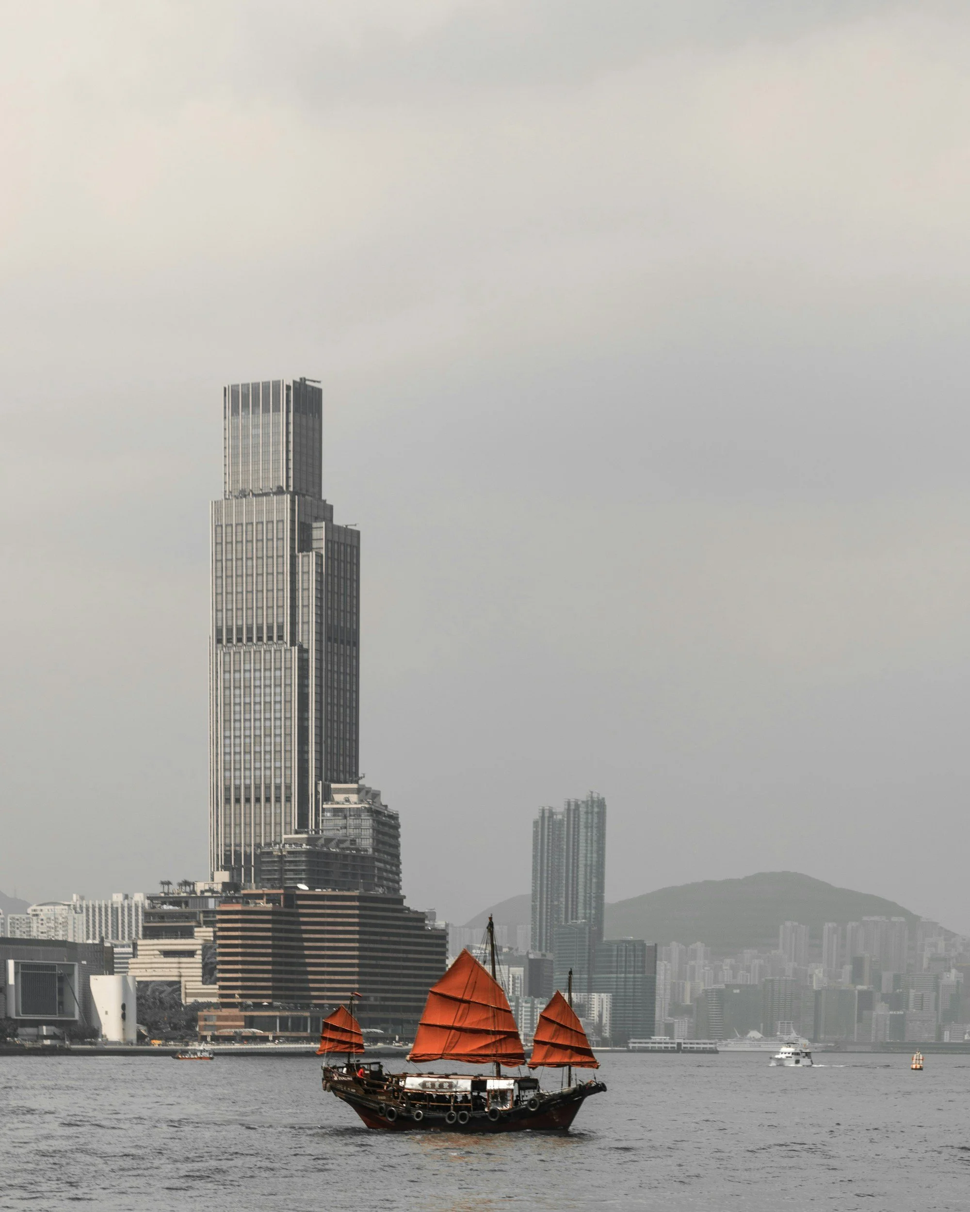 A traditional Chinese junk boat with orange sails sailing on a river in front of a modern city skyline with tall skyscrapers, under an overcast sky.