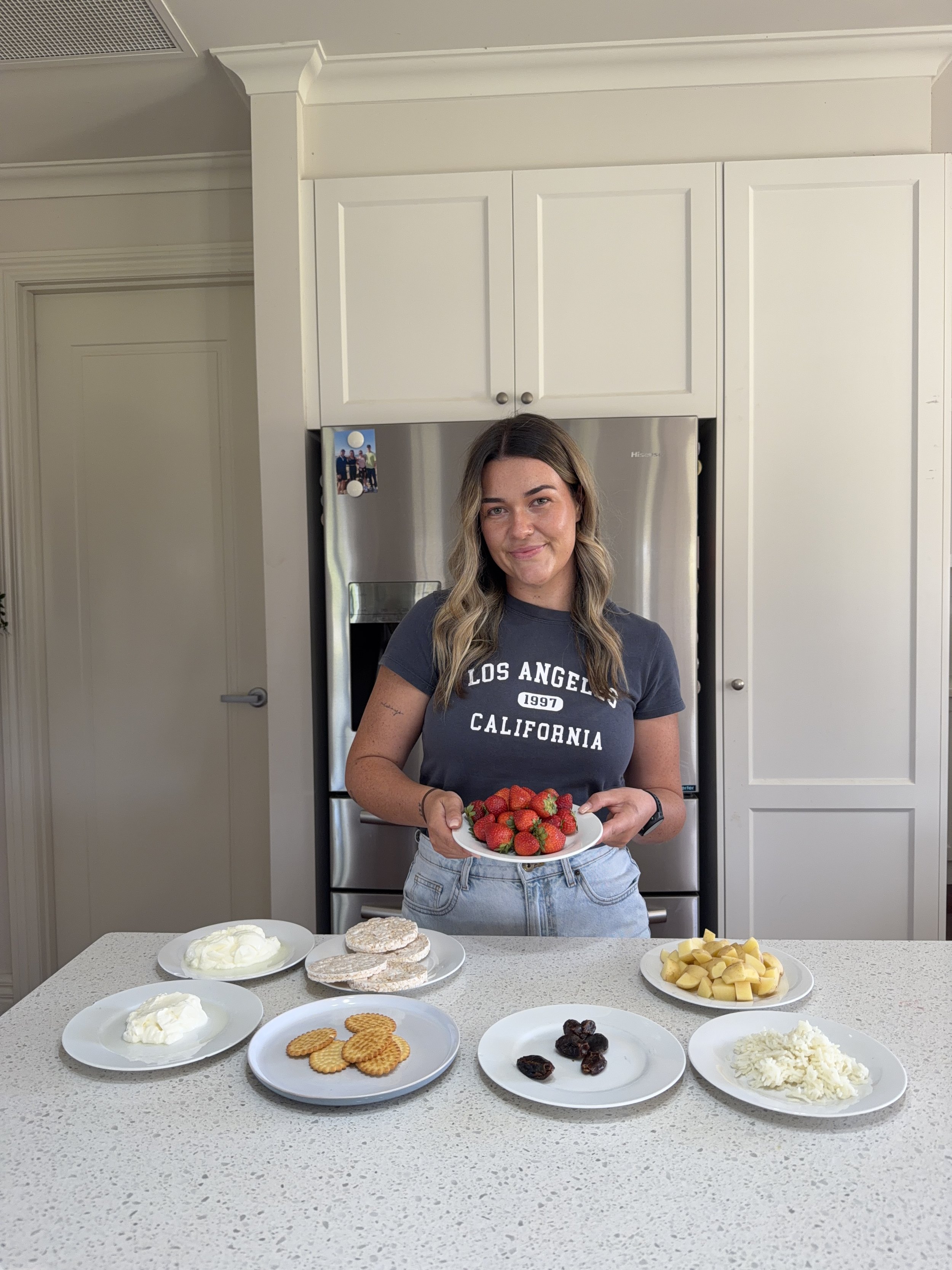 A woman holding a plate of strawberries in a kitchen with a counter with various desserts, including whipped cream, cookies, and fruit, and a refrigerator in the background.
