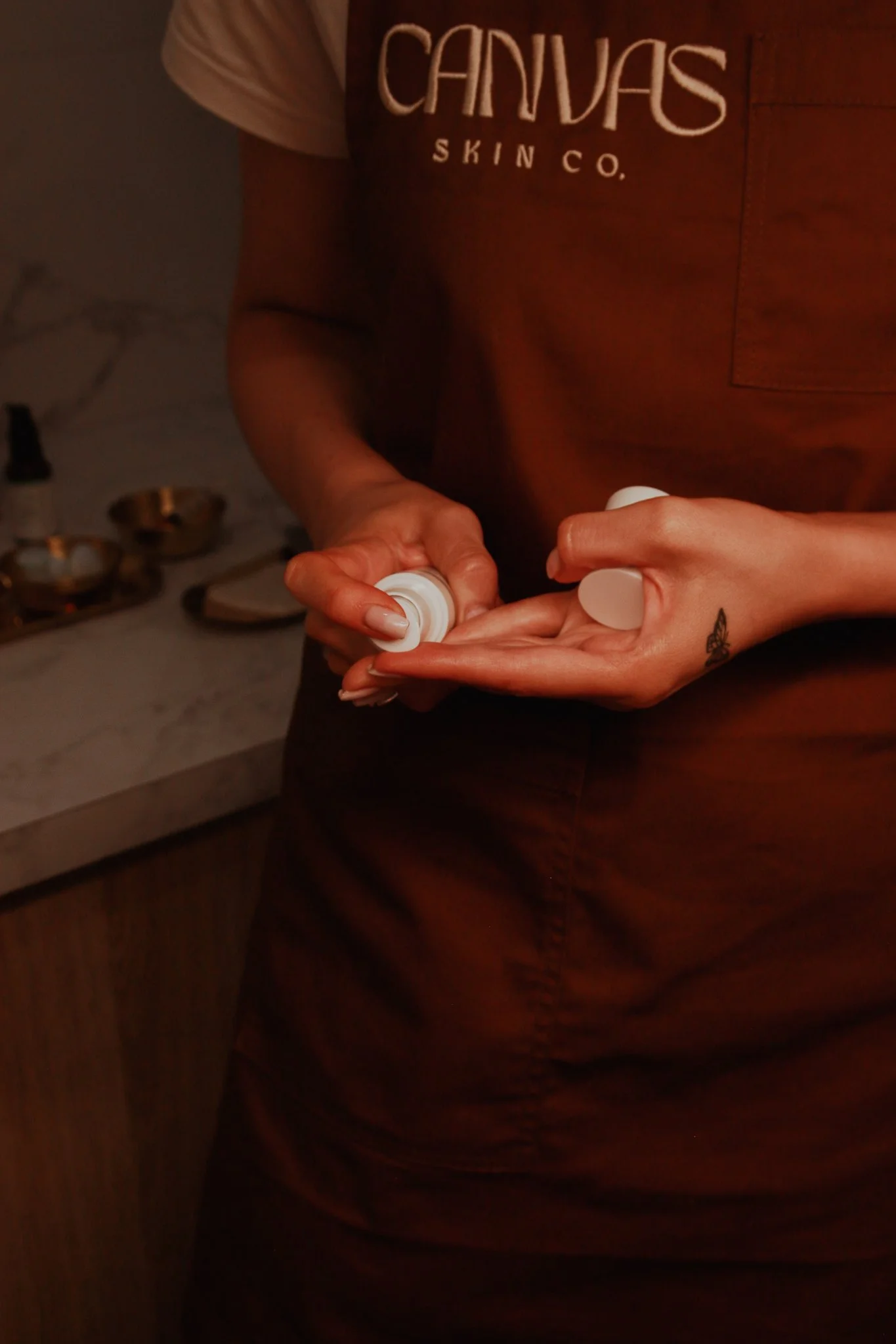 Person wearing a brown shirt with the words 'Canvus Skin Co.' holding a white object, with a tattoo of a butterfly on their wrist, in a dimly lit room with bowls and bottles in the background.