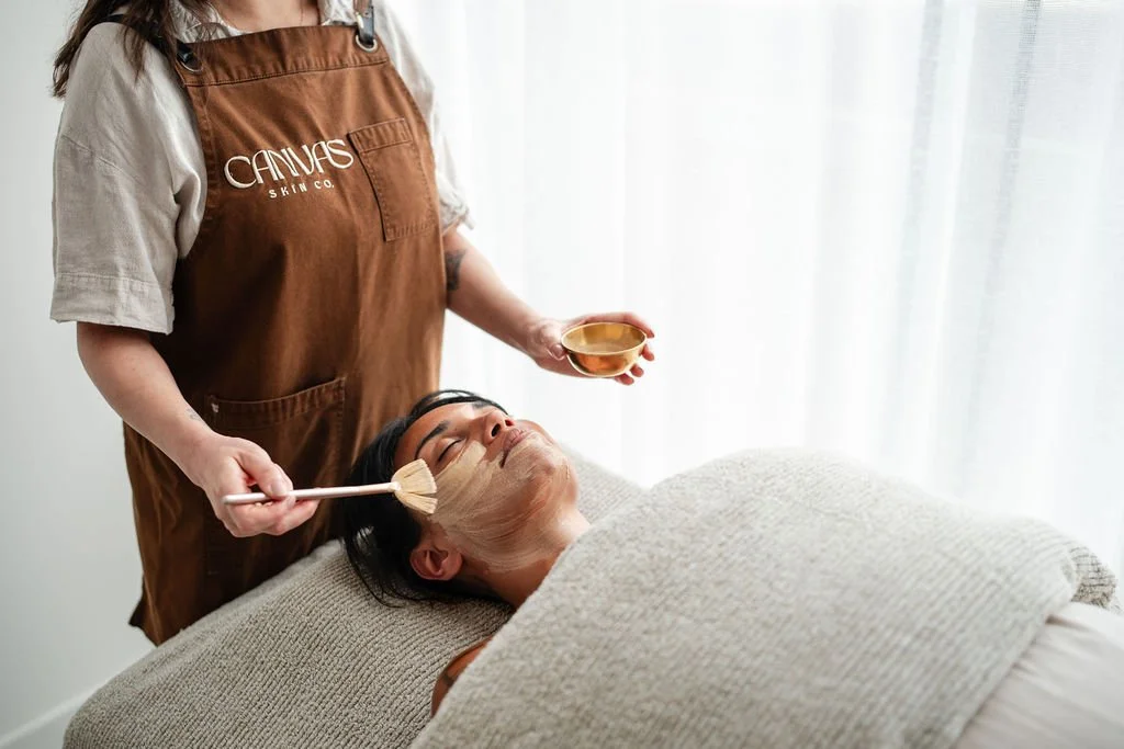 A woman receiving a facial treatment while lying on a massage table, with a beautician applying a facial mask using a brush.