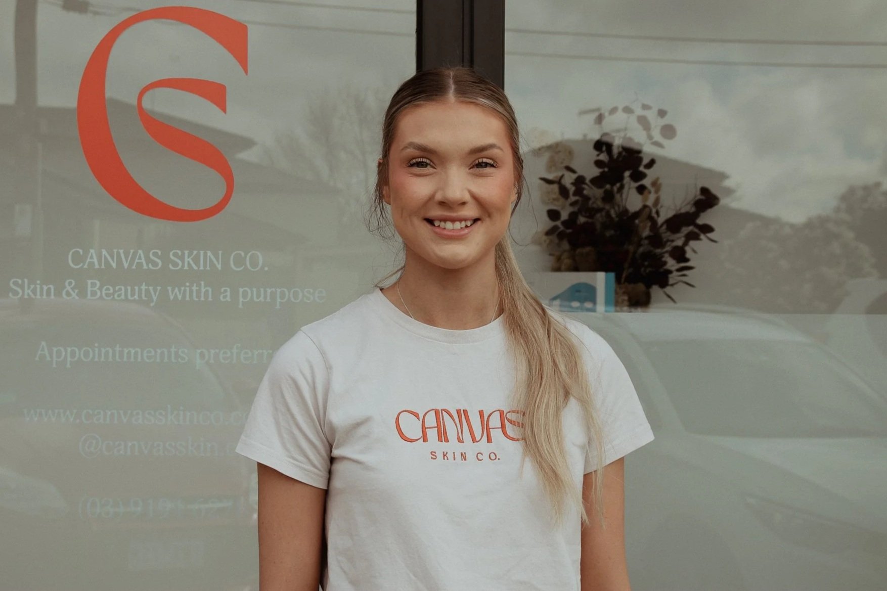 A woman standing in front of a glass door with the logo and name of Canvas Skin Co. on it, smiling at the camera. She is wearing a white t-shirt with the company's logo.
