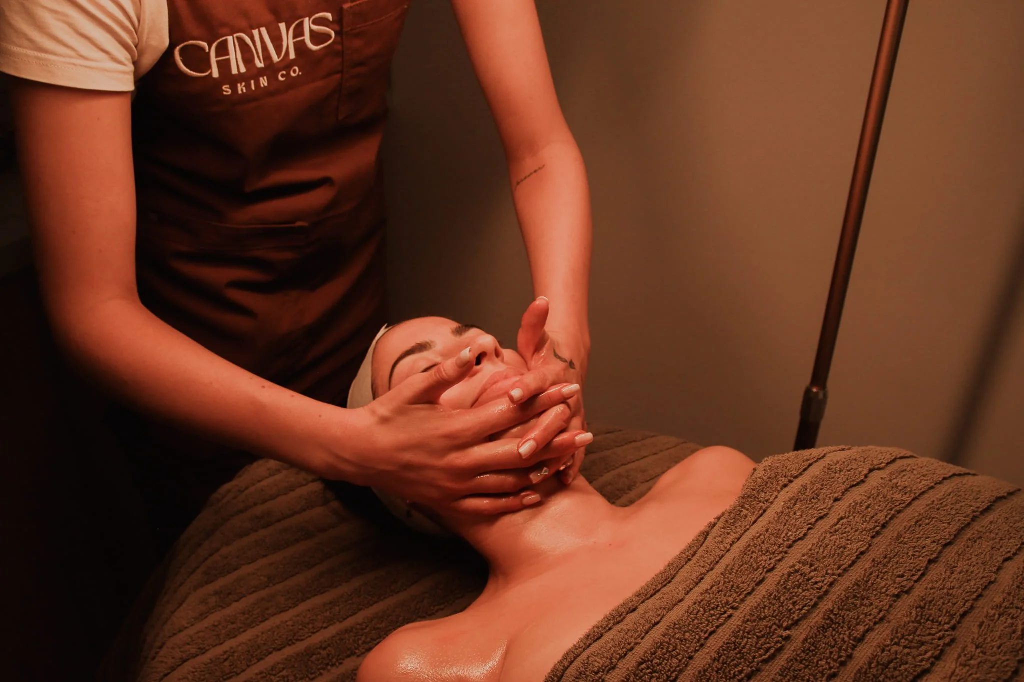 A person receiving a facial massage in a spa setting, lying on a towel with eyes closed, while a therapist applies gentle pressure to their face.