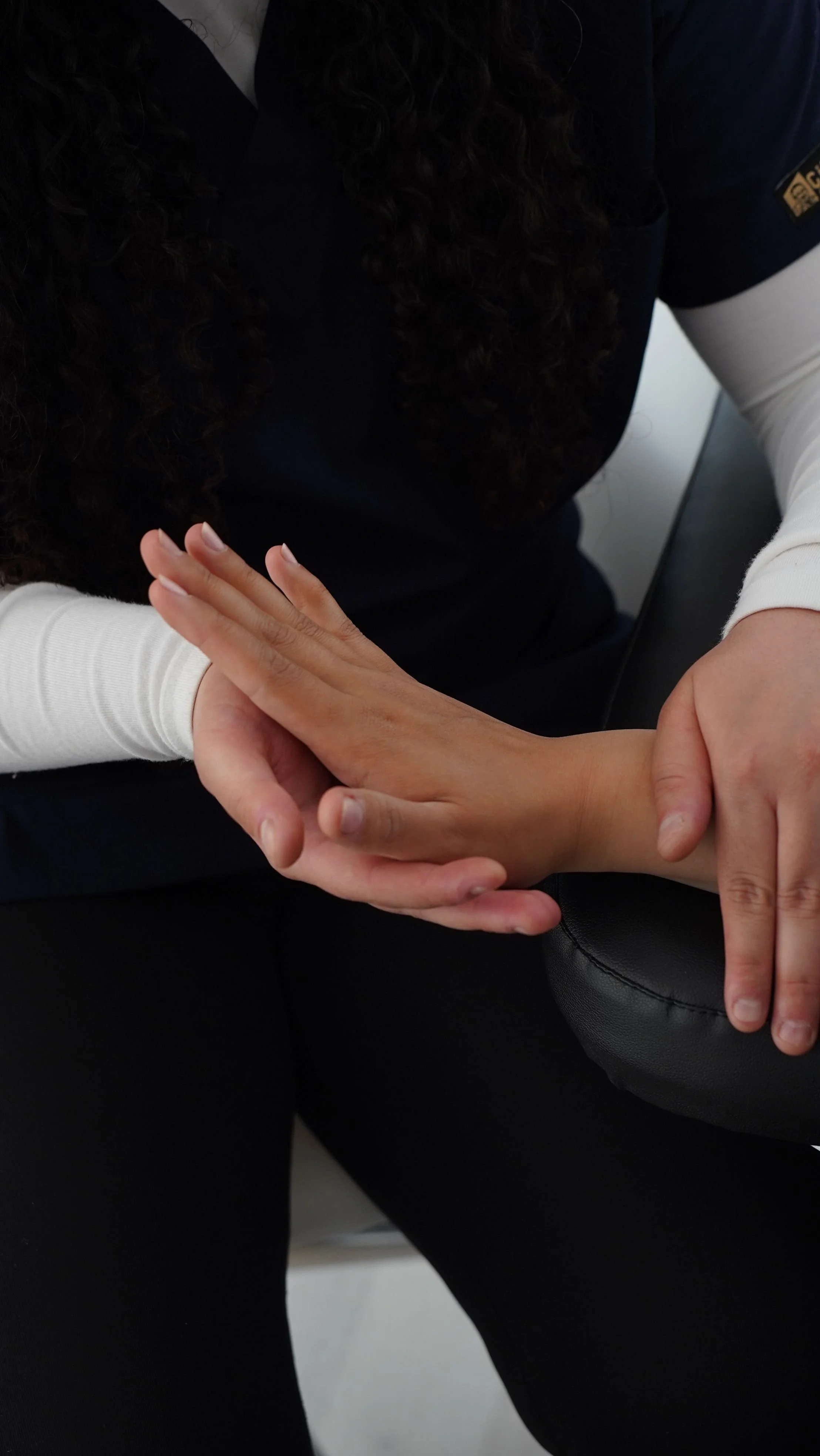 A person with curly dark hair wearing a navy uniform and white long-sleeve shirt receiving a hand massage or handshake from another person.