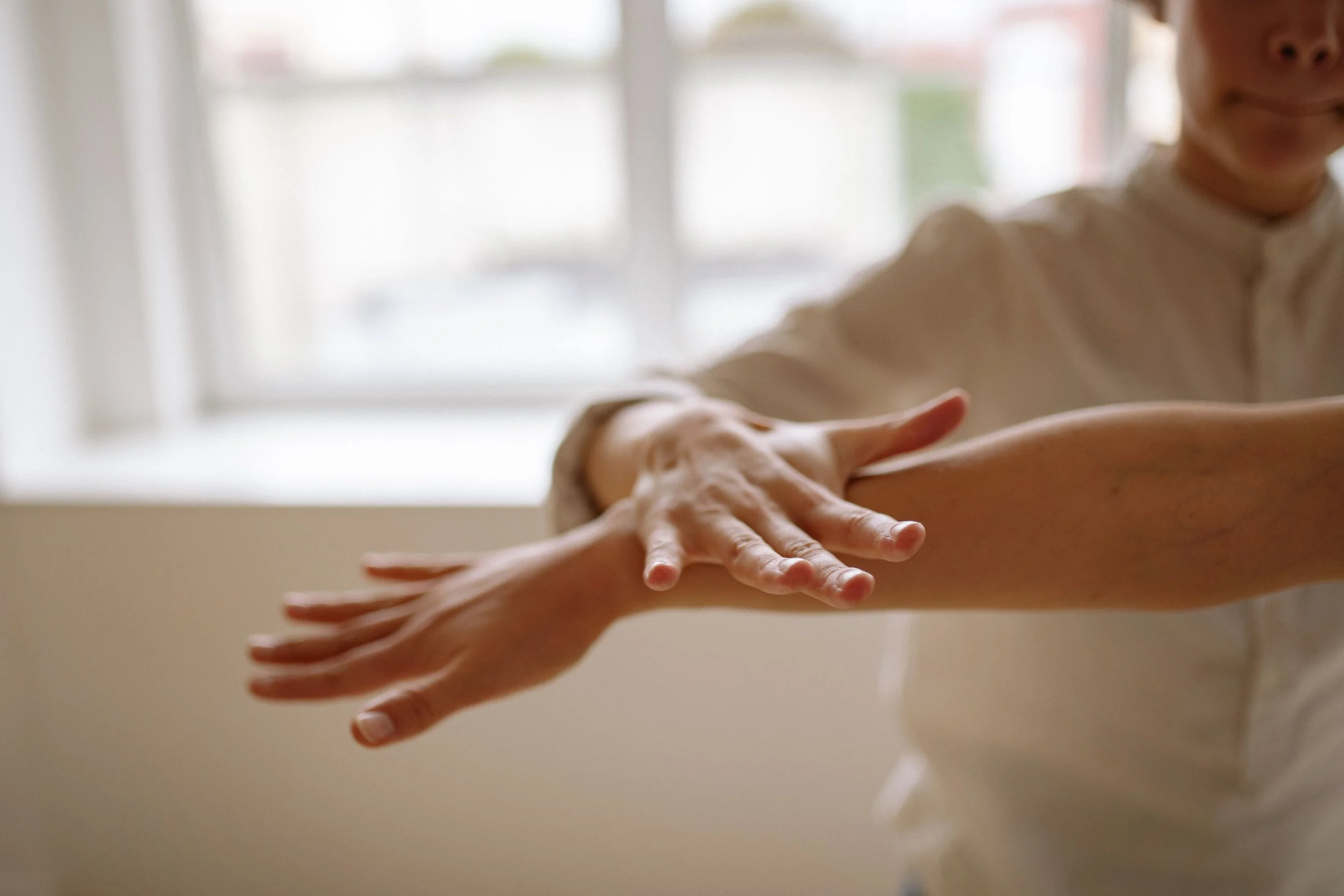 Close-up of two hands, one reaching out and the other resting on the arm of a person, with a blurred background of a window and a person with a neutral expression.