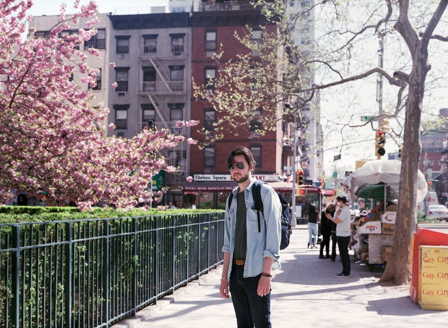 Un jeune homme avec des cheveux foncés, portant des lunettes de soleil, une chemise décontractée, un t-shirt gris et un sac à dos, se tient sur un trottoir dans une rue urbaine avec des arbres en fleurs et des bâtiments en arrière-plan.