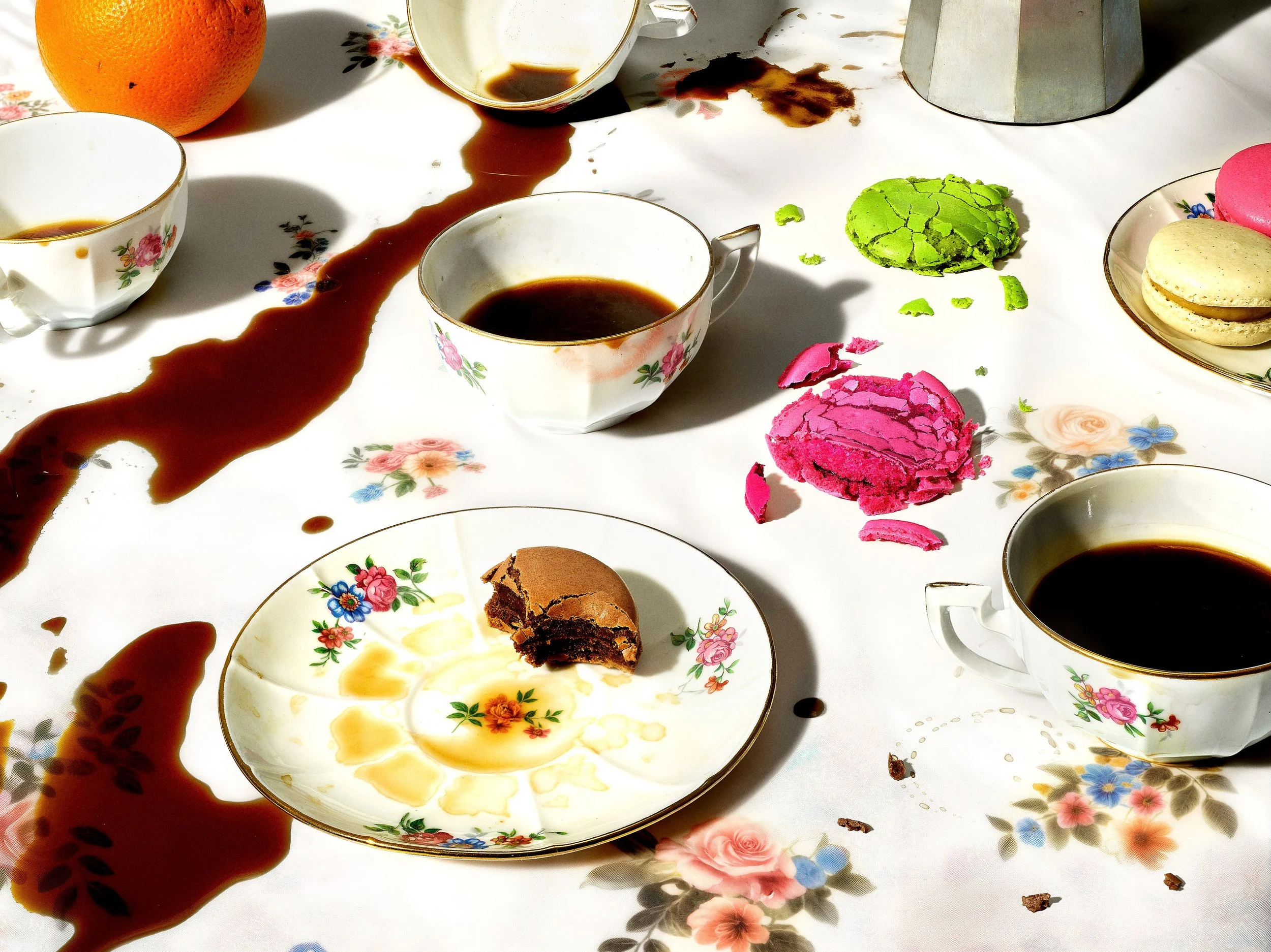 Une table avec des tasses de thé, des macarons, un morceau de gâteau chocolat, des éclats de macarons colorés, une orange et une cafetière, avec du sirop de chocolat renversé sur la nappe.