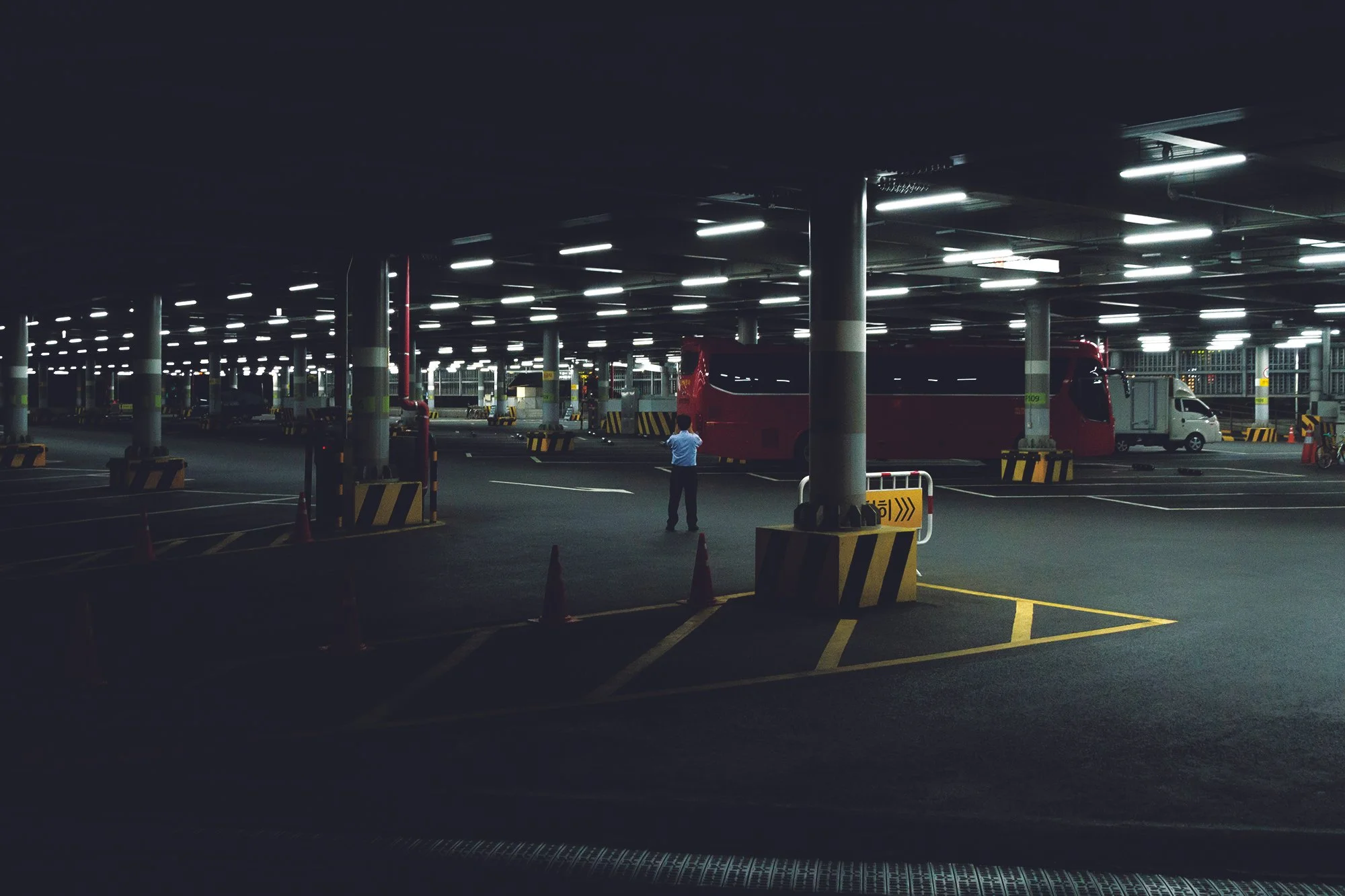 Parking souterrain vide avec des voitures, un autobus rouge, un minibus blanc, et un homme en train de prendre une photo, éclairage fluorescent au plafond.