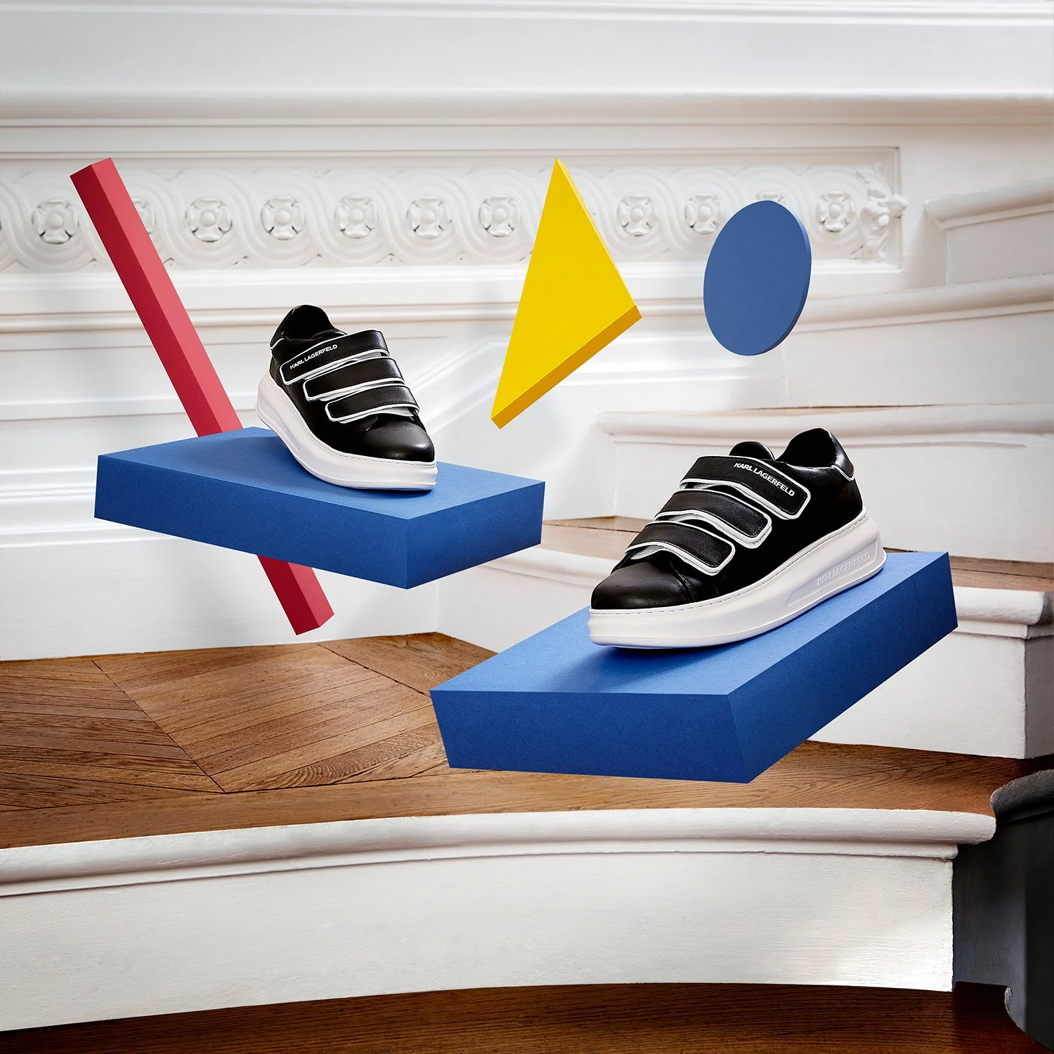 Deux baskets à talons noirs avec bandes, posées sur des socles bleus flottants dans une pièce au mur blanc avec moulures décoratives.