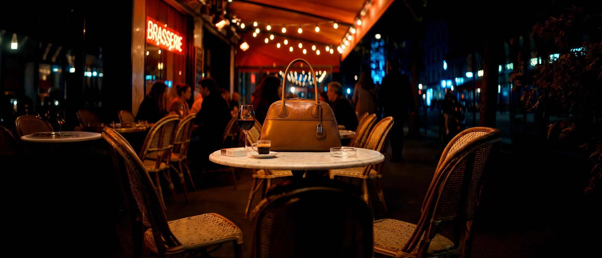 Une scène de terrasse de café ou restaurant la nuit, avec une table au centre portant un sac à main beige, deux verres de vin rouge, une cigarette, une boîte de cigarettes et un cadenas. En arrière-plan, des personnes discutent sous une enseigne lumineuse 'Brasserie' avec des guirlandes lumineuses suspendues.