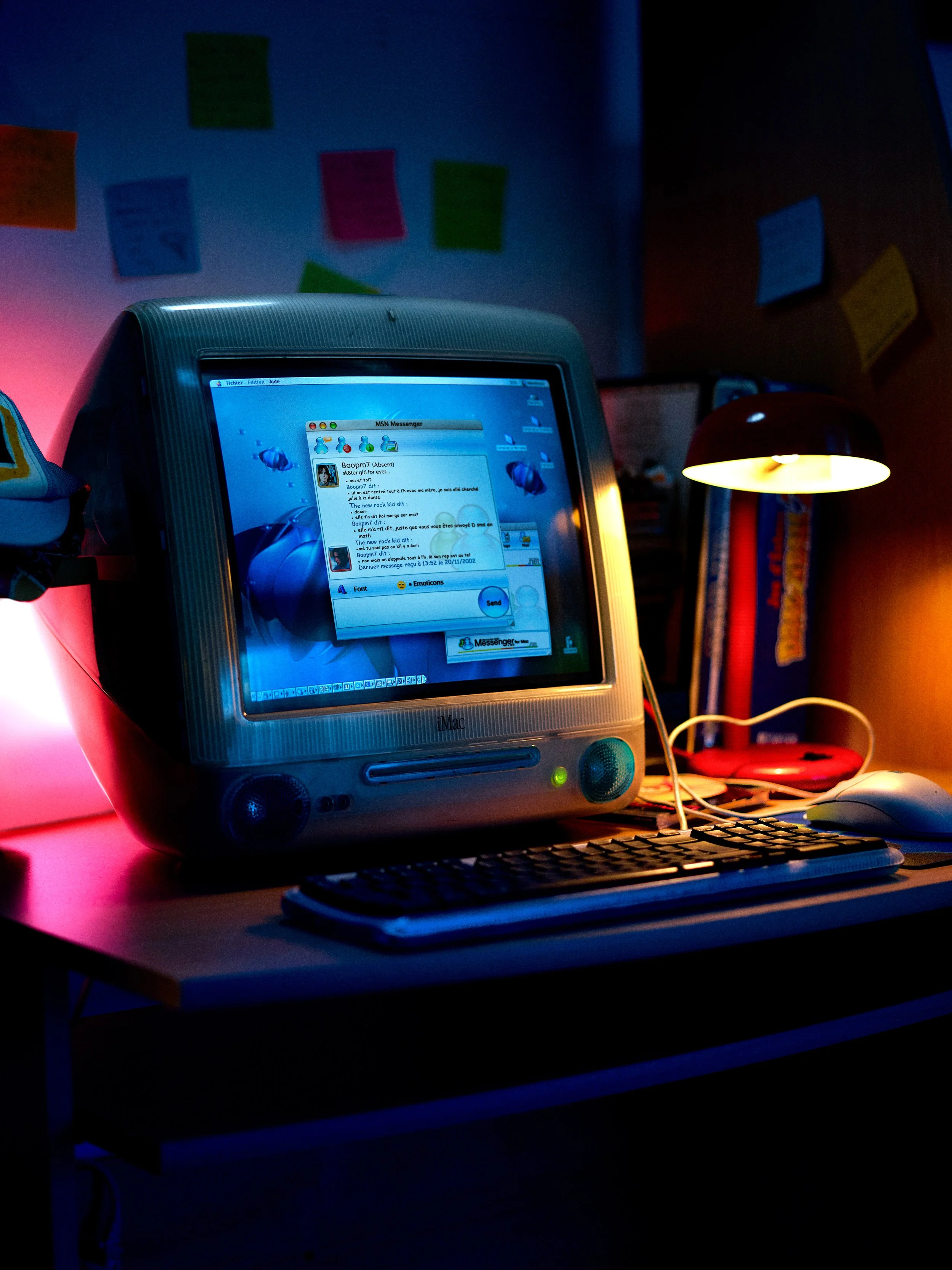 Un ordinateur vintage avec un écran d'iMac affichant une messagerie instantanée, placé sur un bureau avec clavier et souris. En arrière-plan, une lampe rouge et un mur décoré de Post-it colorés dans une pièce sombre.