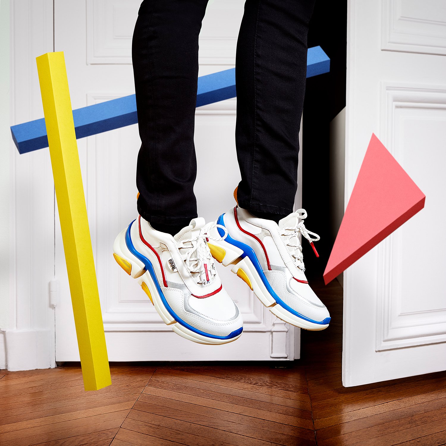 Chaussures de sport blanches avec des accents de bleu, rouge et jaune, suspendues dans une pièce, encadrées par des formes géométriques colorées