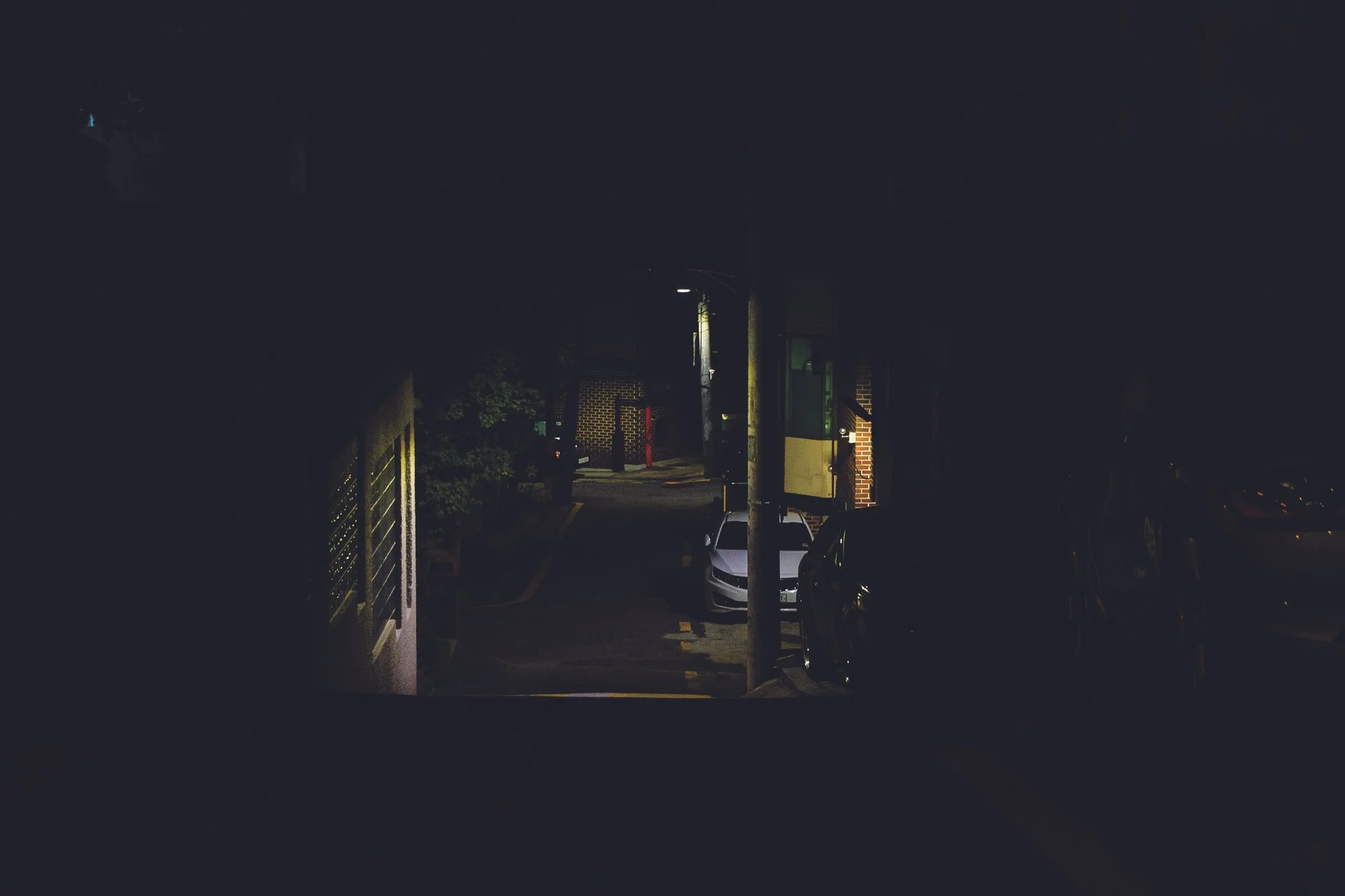Une rue sombre avec un seul éclairage public, des voitures garées et un bâtiment en arrière-plan, vue la nuit.