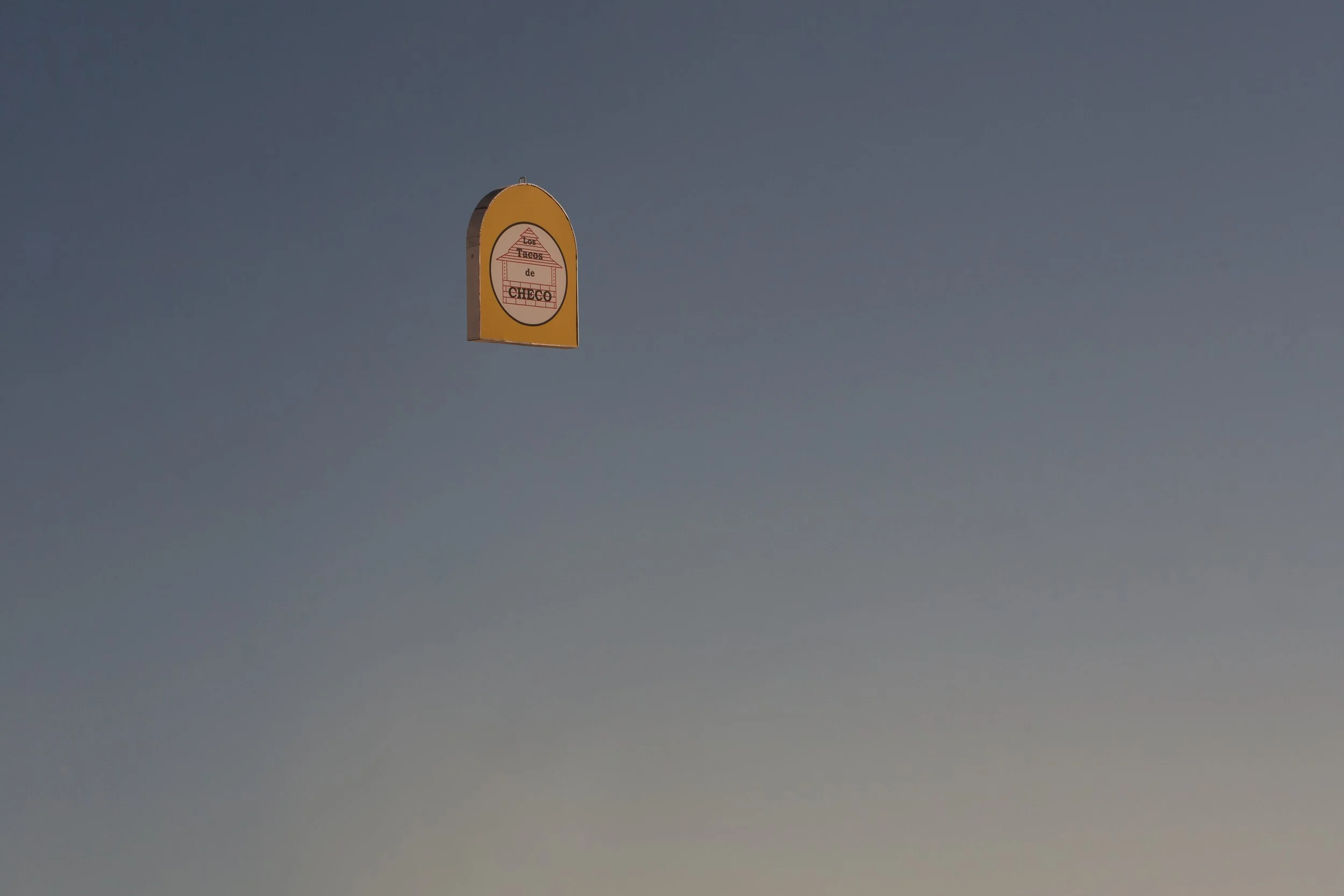 Un panneau de signalisation avec un fond jaune et un logo circulaire représentant une maison de tacos de couleur rouge avec un toit en tuiles, inscrit avec le texte "Tacos de Checo".