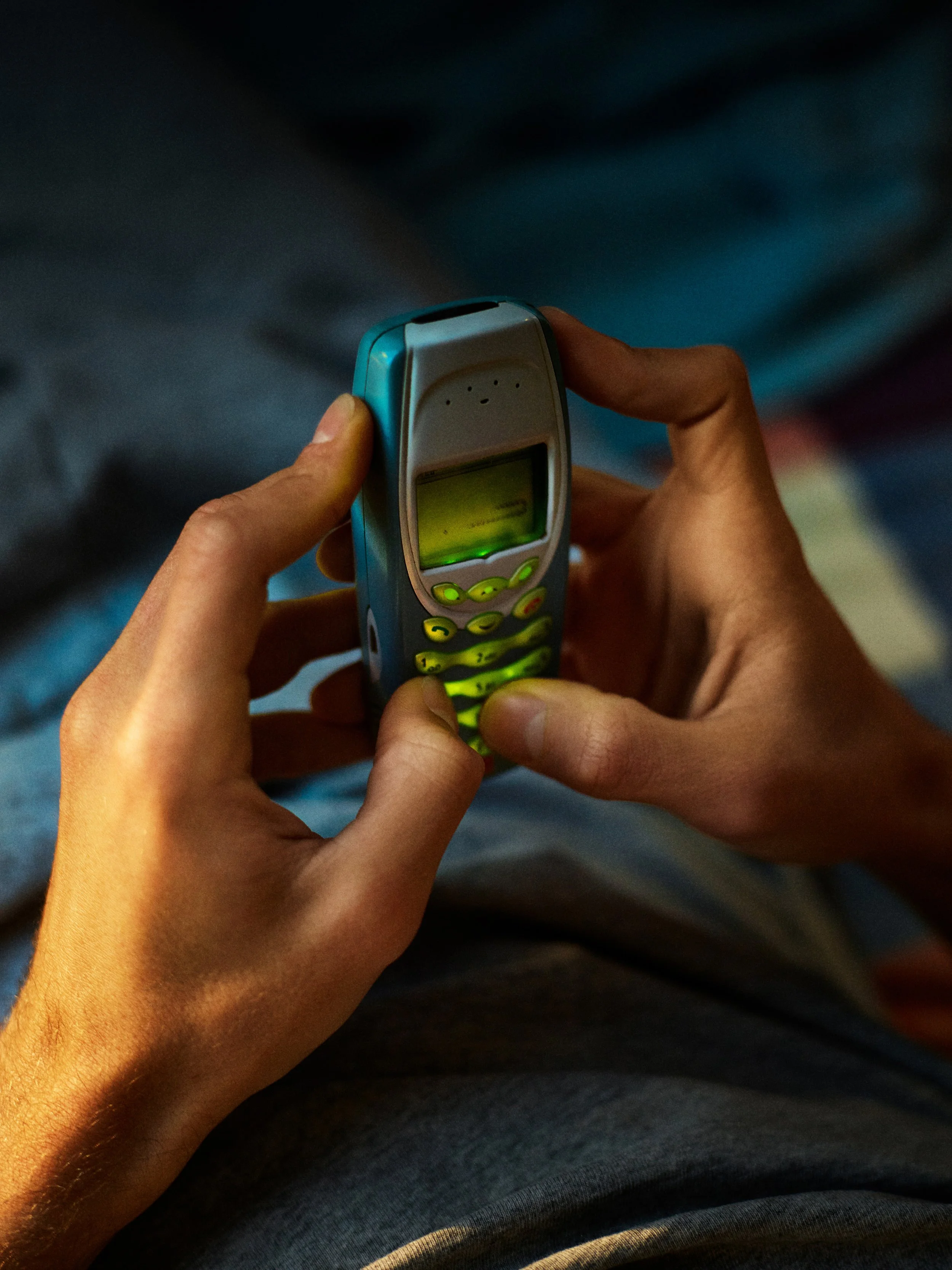 Une personne tient un ancien téléphone portable en main, avec un écran allumé. La photo est prise par-dessus, et la personne porte un vêtement sombre.
