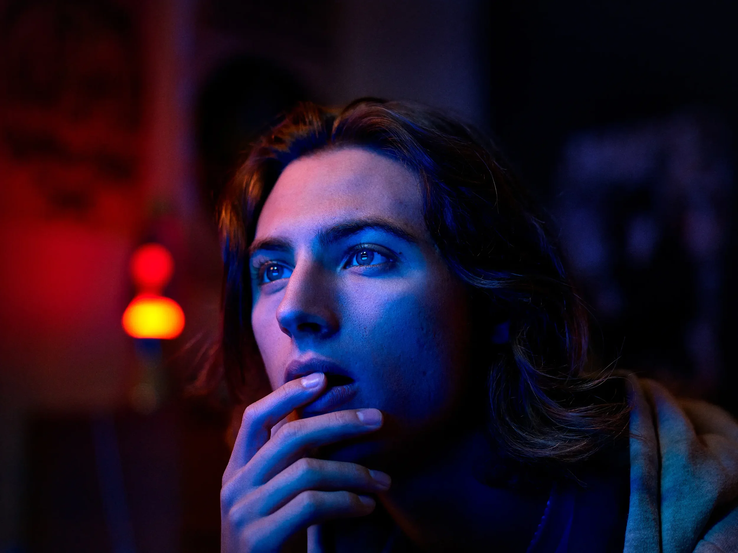 Une jeune femme aux yeux bleus regarde pensivement dans l'obscurité avec un doigt posé près de sa bouche, éclairée par une lumière colorée bleue et rouge.