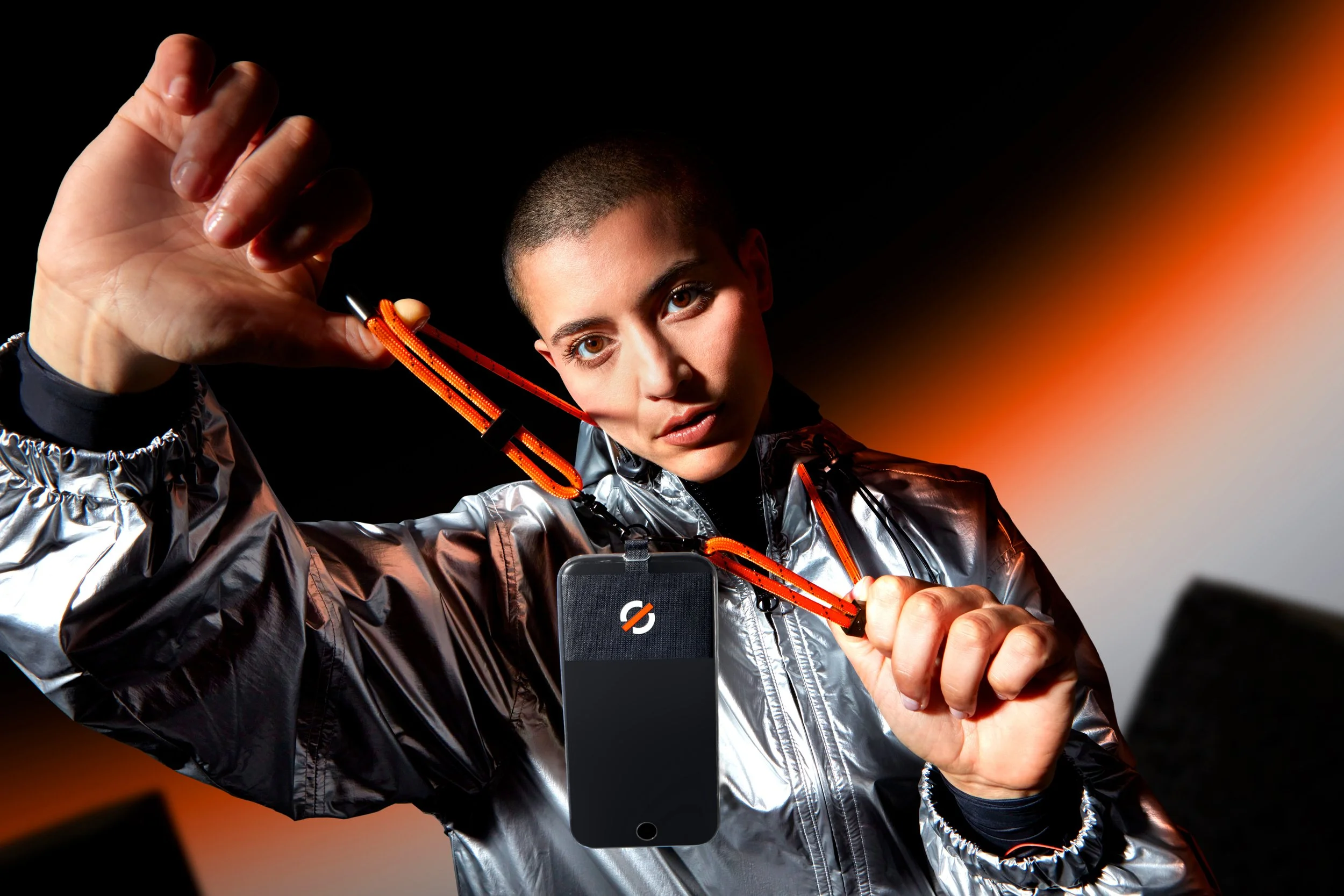Une personne avec une tête rasée, portant une veste argentée, tient un smartphone en montrant un badge ou une carte avec un symbole barré. Elle se prépare à prendre une photo dans un environnement sombre avec une lumière orange diffuse.