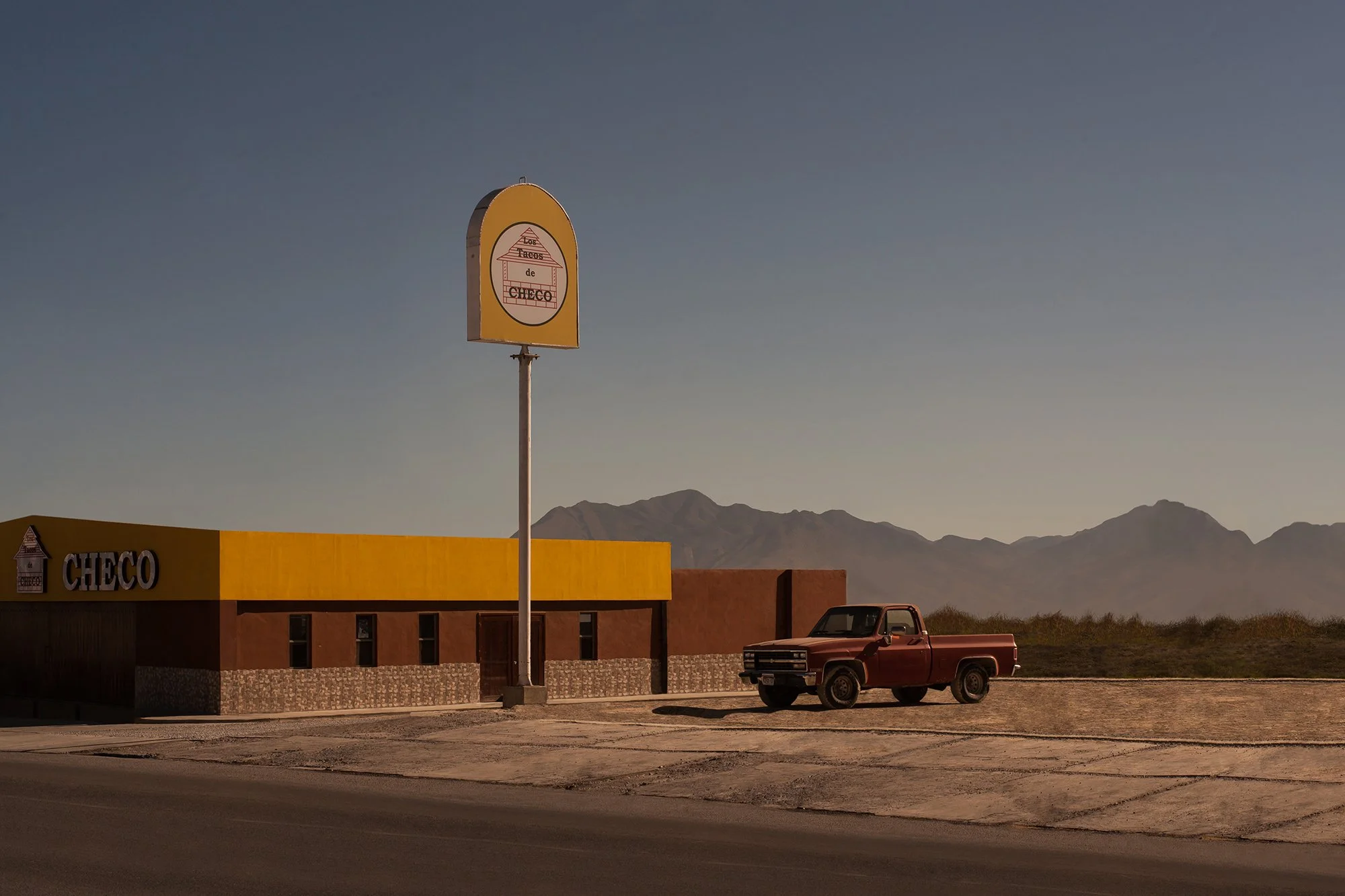 Une station-essence jaune et un camion rouge garé devant, avec des montagnes en arrière-plan.
