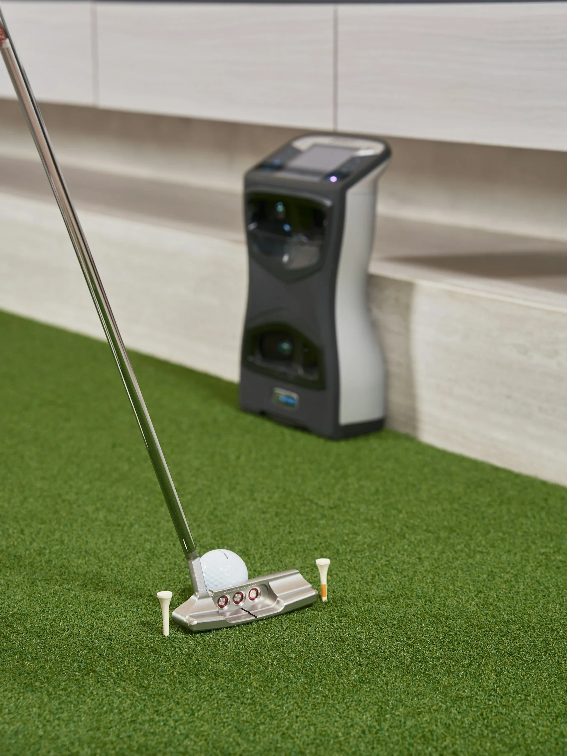 Close-up of a golf putting setup on a synthetic grass surface, with a golf ball, tee markers, and a putter in focus, and a golf swing game machine and light-colored wall in the background.