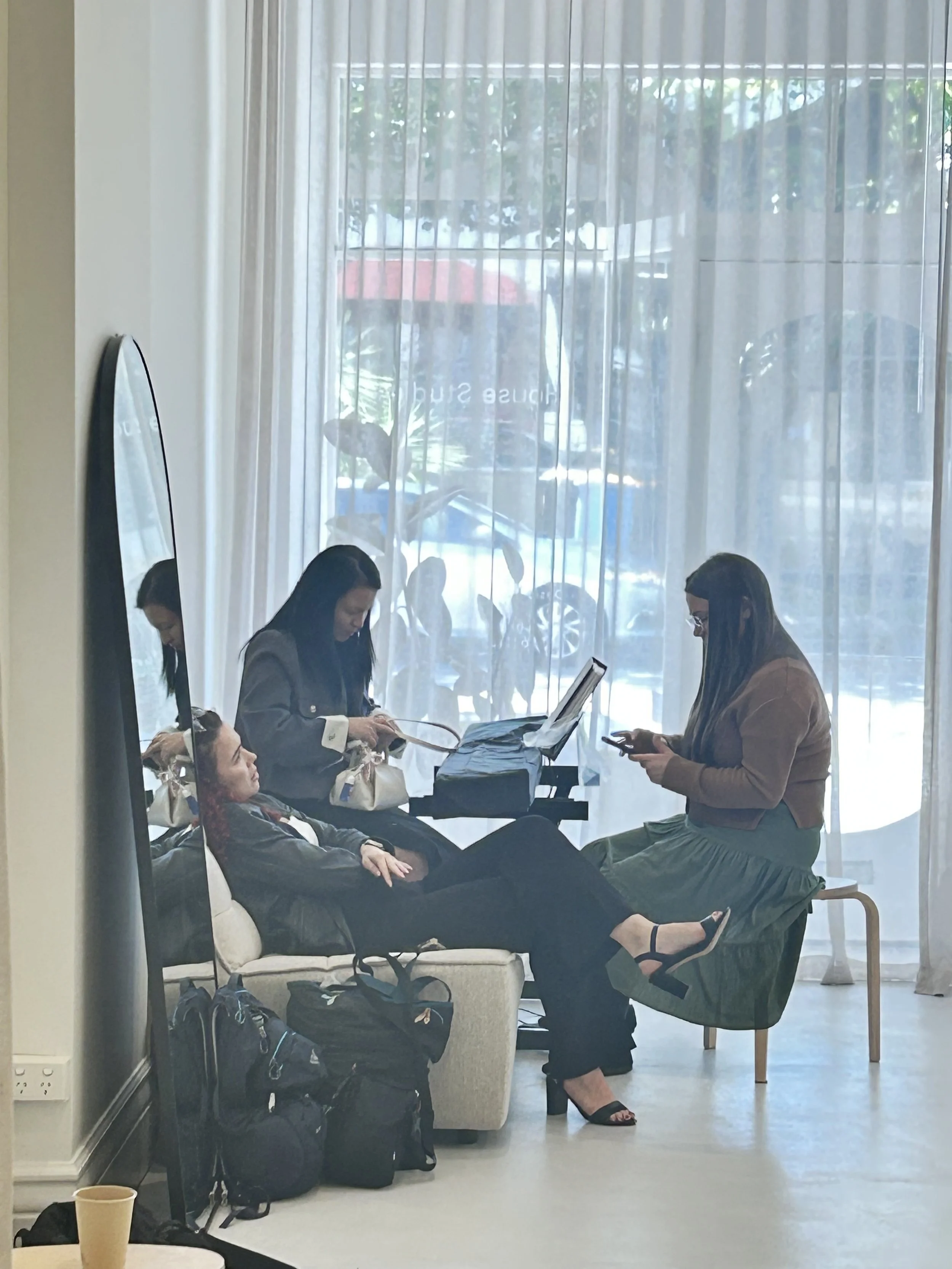 Three women in an indoor waiting area, with one seated and two standing, all using their phones. Bags are on the floor and a window with sheer curtains is in the background.