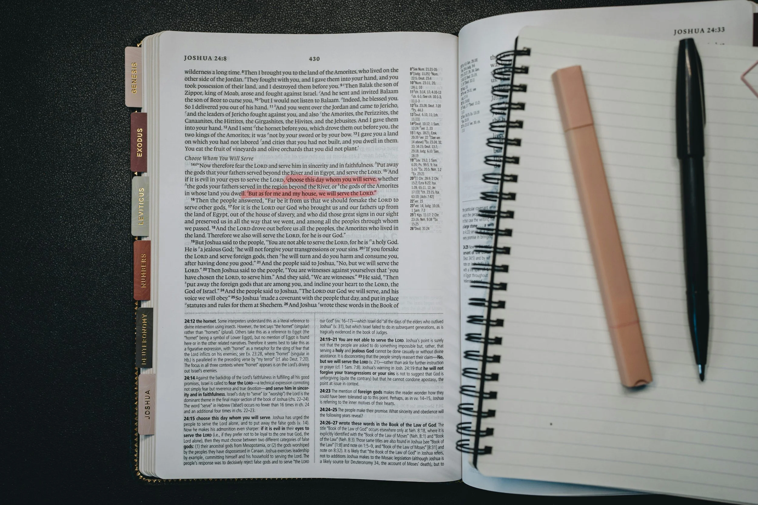 Open Bible with tabs on the side showing books Genesis, Exodus, Leviticus, Numbers, and Deuteronomy. The page displayed is Joshua 24:8 to 26:27, with the book header and chapter details visible. A black pen and a pink sticky note are clipped on the right page, resting on a spiral notebook.