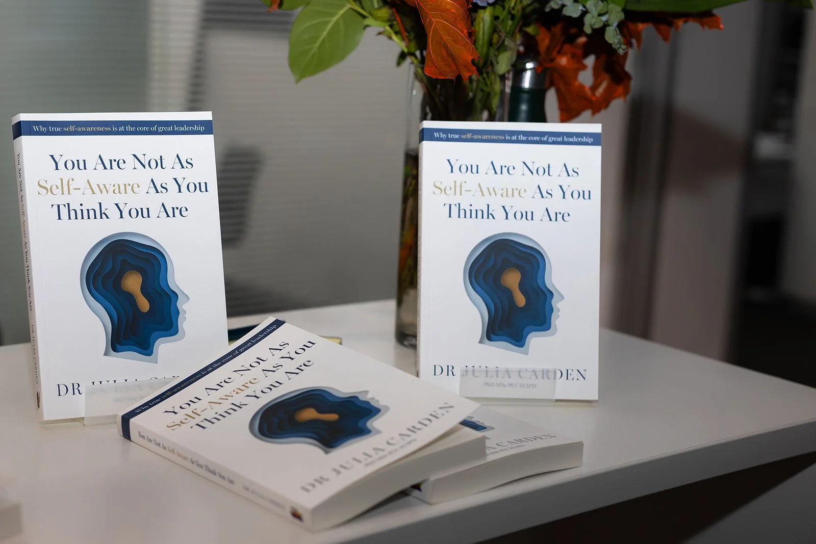 Three copies of a book titled 'You Are Not As Self-Aware As You Think You Are' by Dr. Julia Gorden are displayed on a white table. One book stands upright behind a potted plant, one is upright next to the plant, and one is lying flat in front. The plant has green and reddish leaves, and the background includes a blurred gray wall and shelves.