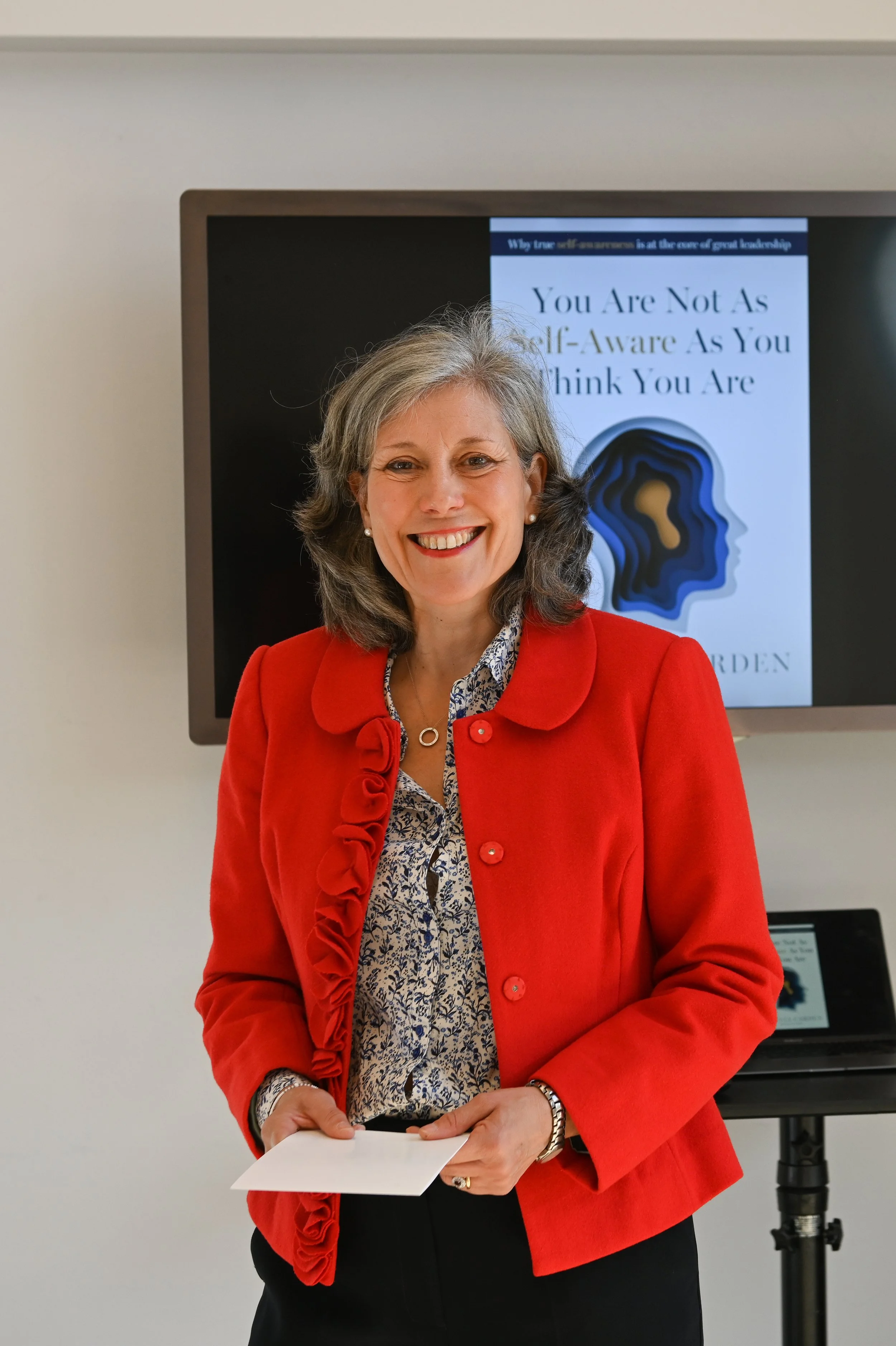 A woman with gray hair and pearl earrings, wearing a red blazer with ruffle details, standing in front of a presentation screen with a slide that reads 'You Are Not As Self-Aware As You Think You Are'. She is smiling and holding a piece of paper.