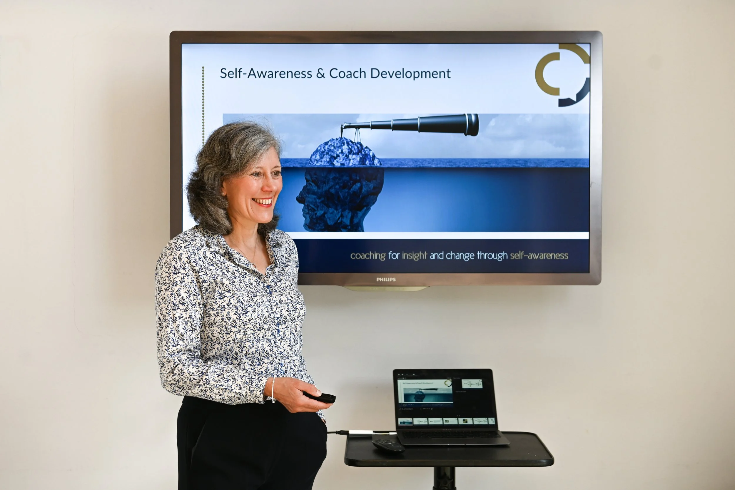 A woman with gray hair smiling and holding a remote control, standing in front of a presentation screen that says 'Self-Awareness & Coach Development' with images of a telescope and a rock in the ocean. There is a small computer or tablet on a stand and a keyboard on a table.