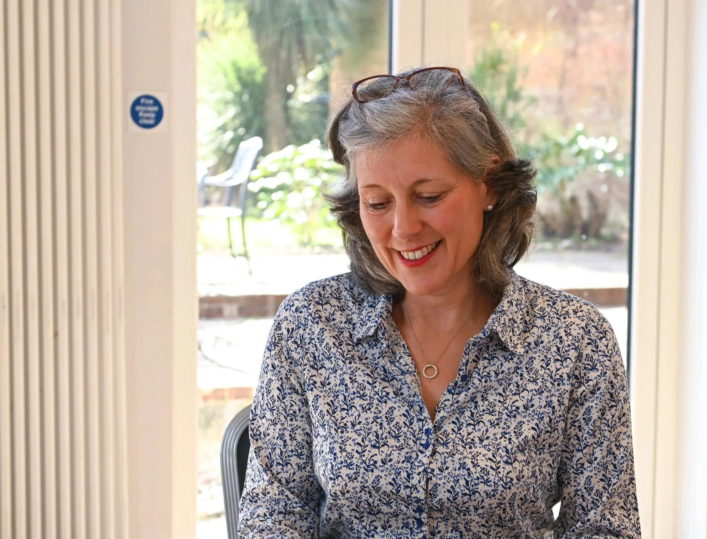 A woman with gray hair and glasses on her head wearing a patterned blouse smiling, sitting in front of a glass door with a garden visible outside.
