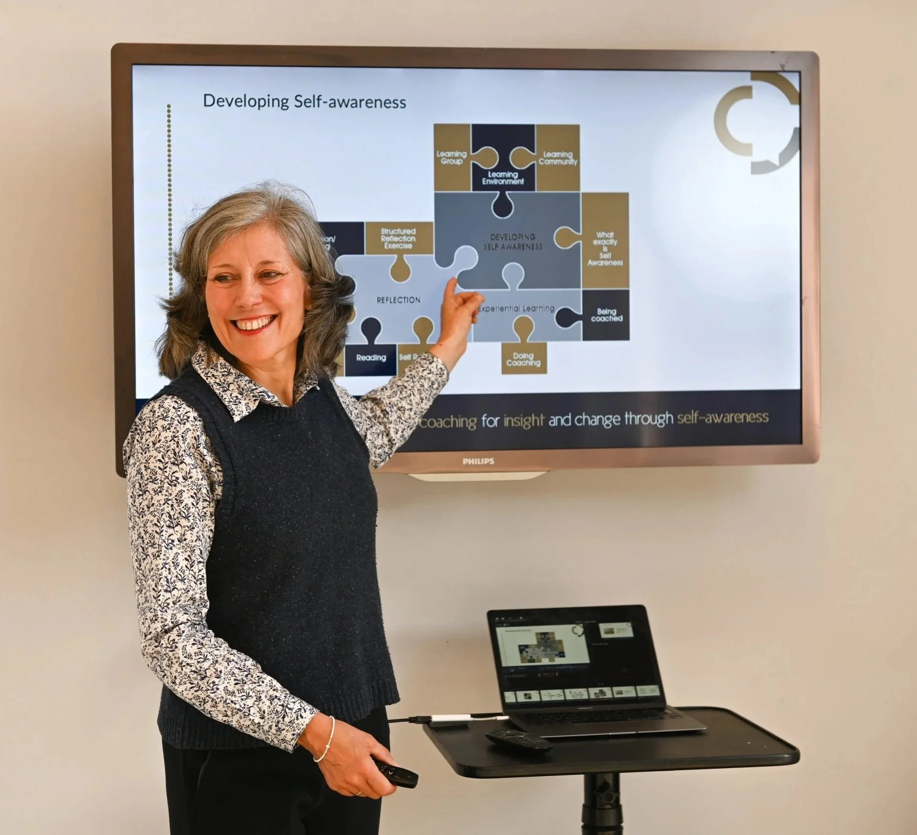 A woman standing in front of a screen presenting a slide titled 'Developing Self-awareness.' She is smiling and pointing to the slide which contains a puzzle diagram with words related to self-awareness and learning. She holds a remote control in her right hand, with a laptop on a stand below the screen.