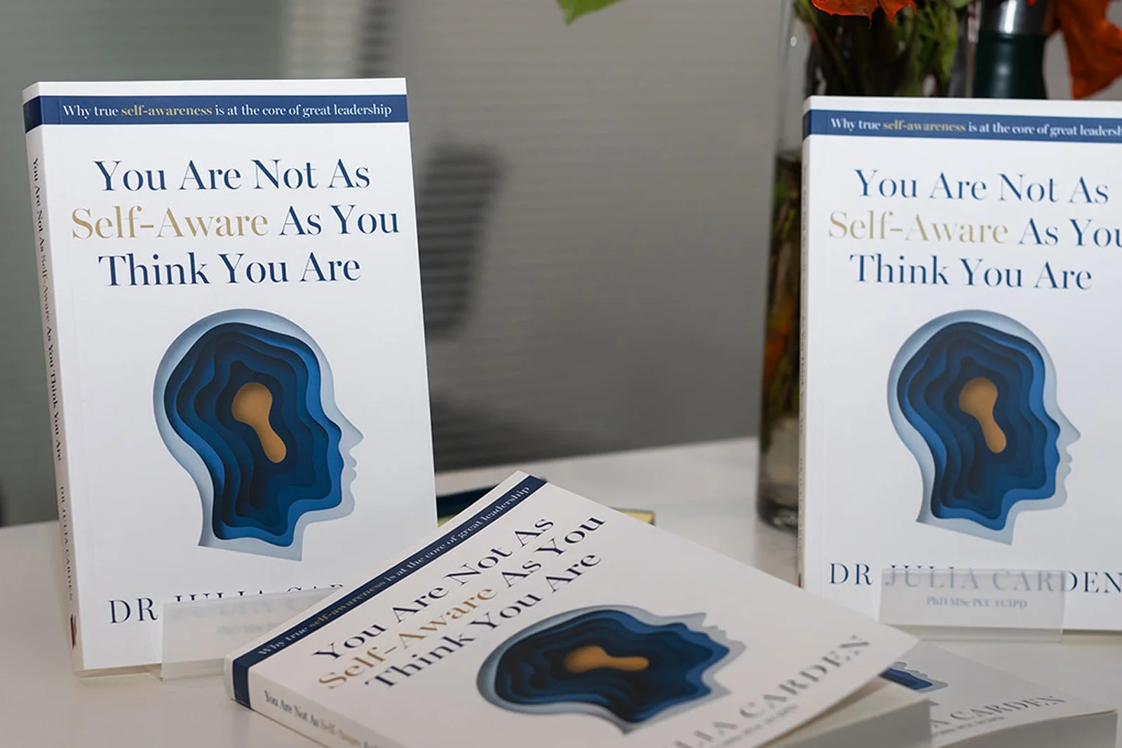 Multiple copies of a book titled 'You Are Not As Self-Aware As You Think You Are' by Dr. Julia Carden are displayed on a table, with some standing upright and others lying down.