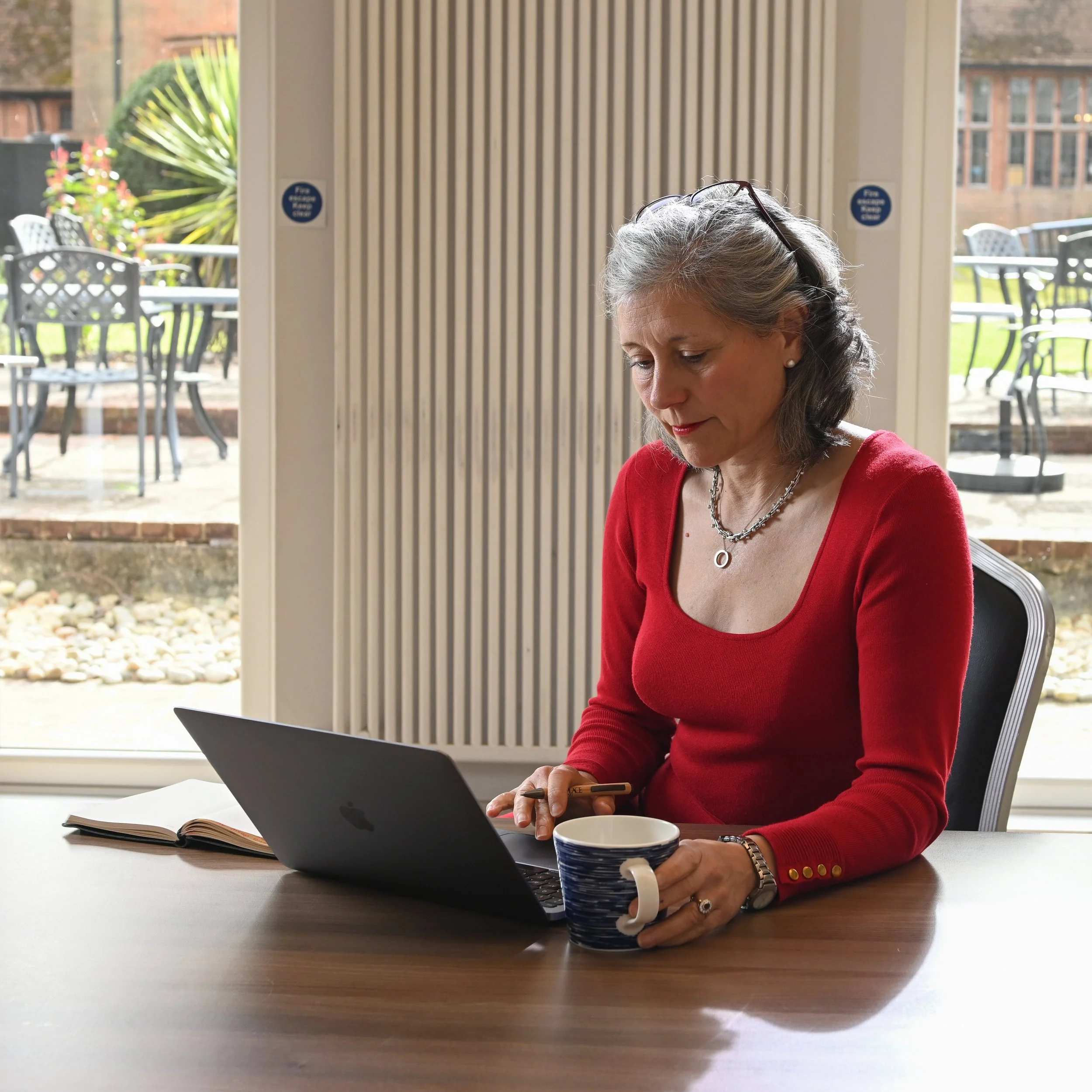A mature woman in a red sweater sitting at a desk with a laptop, reading a phone, with a mug, an open book, and a pen on the desk in a bright interior with large windows and outdoor patio furniture visible in the background.