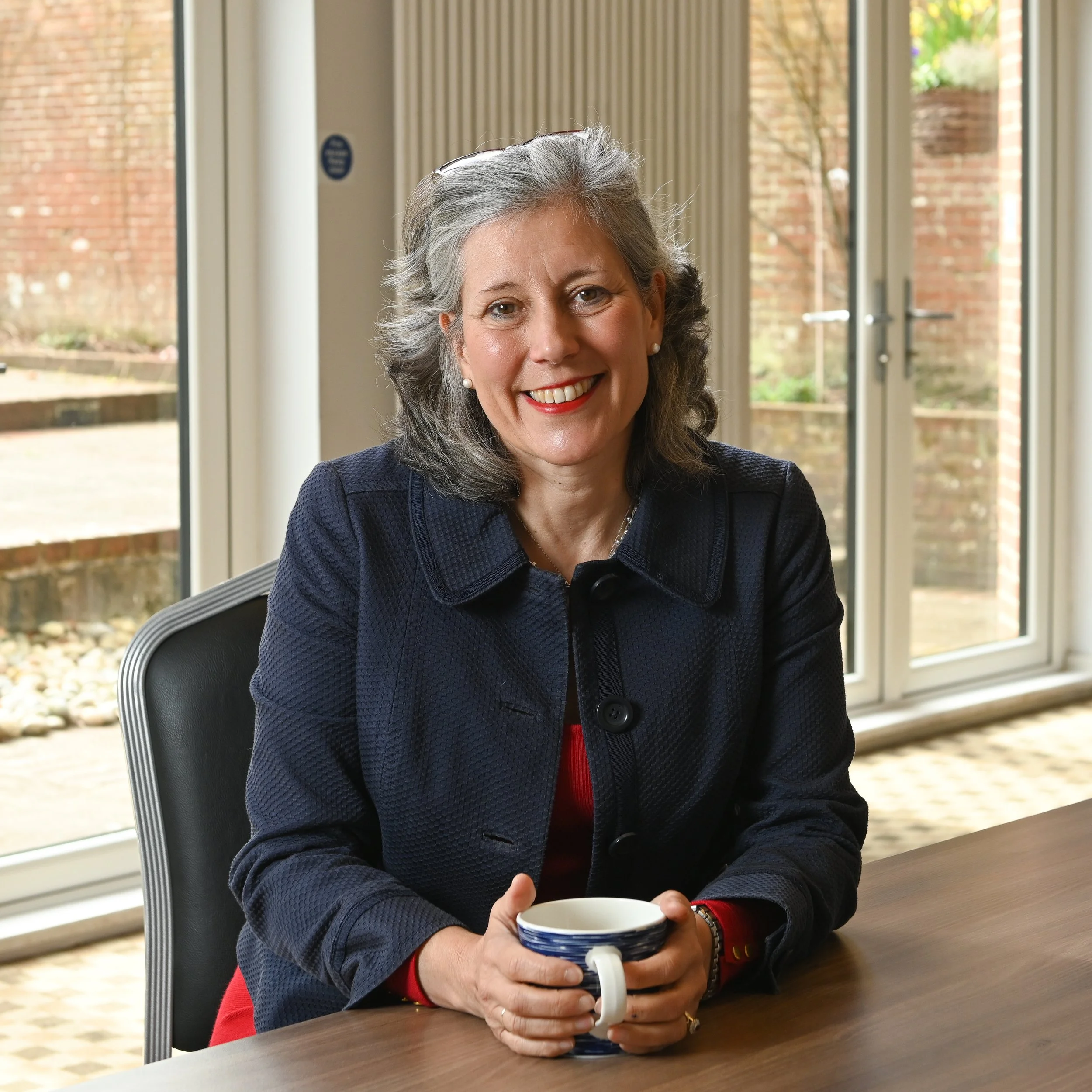 A smiling woman with gray hair and pearl earrings sitting at a table, holding a mug with both hands, in a room with large windows and brick outside view.