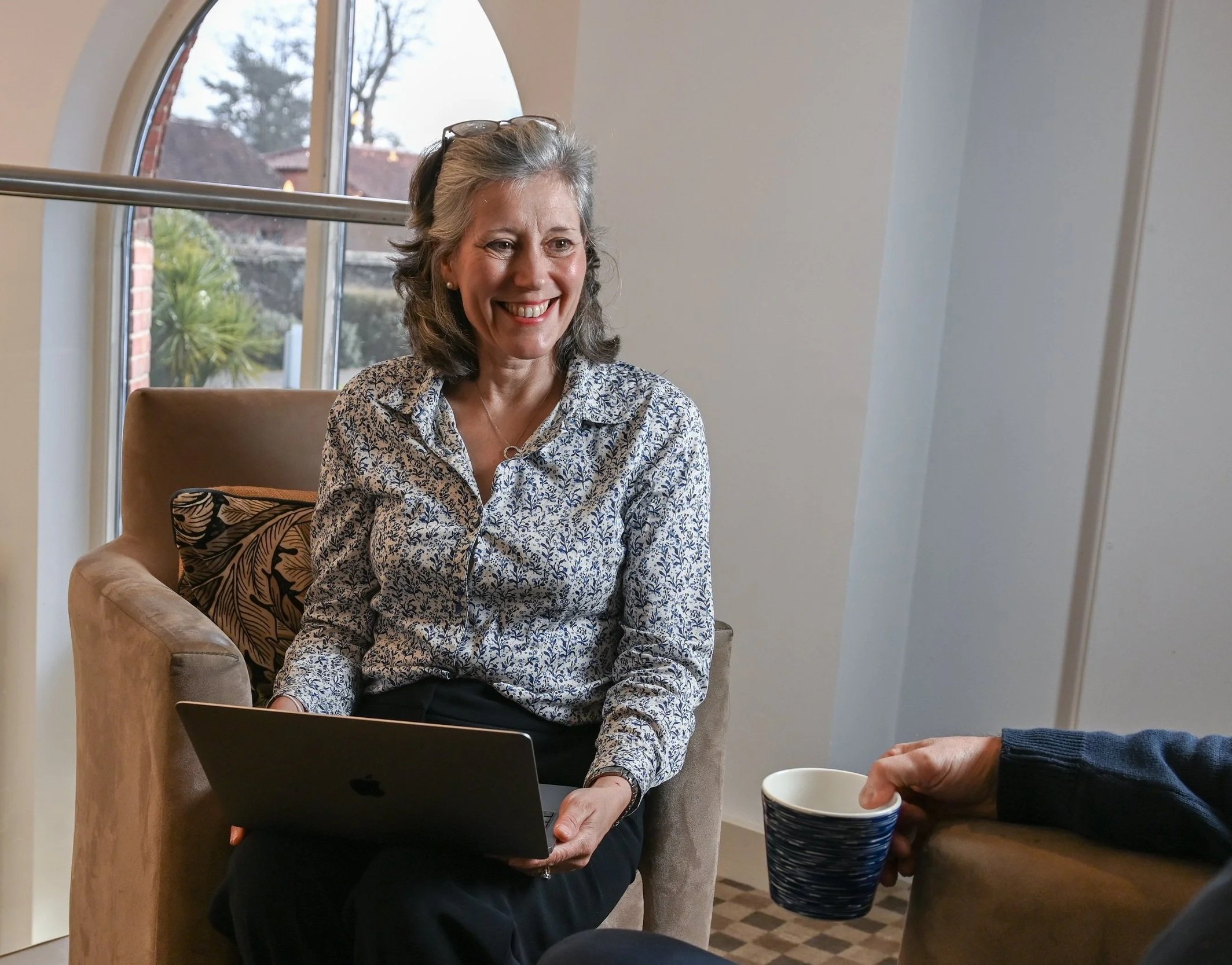 A smiling middle-aged woman with gray hair, wearing glasses on her head, a patterned blouse, and earrings, sitting in a cozy room with light-colored walls and large windows, holding a laptop on her lap. She is engaged in conversation with another person, who is holding a cup, in a warm and inviting environment.
