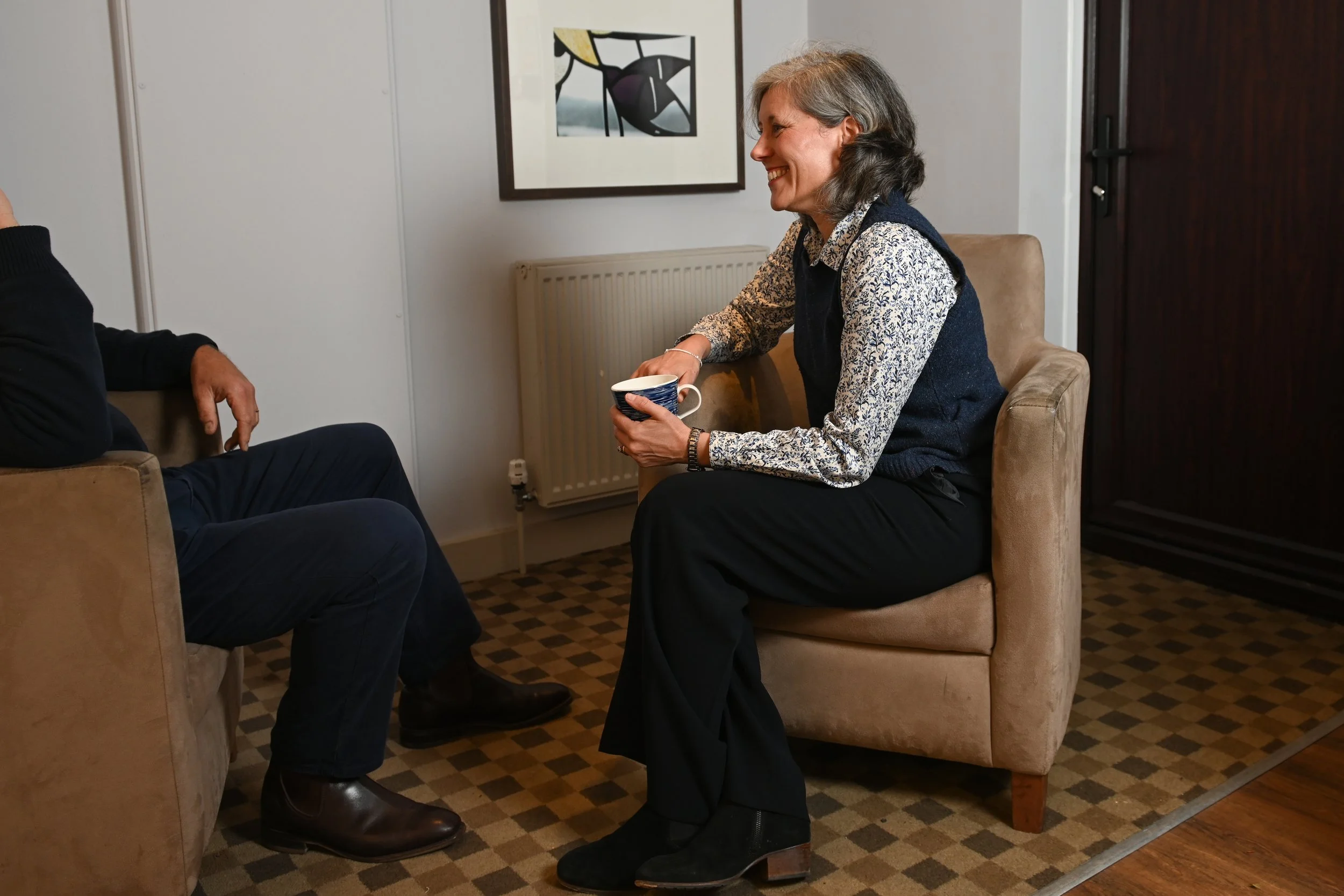 A woman smiling and holding a coffee mug, sitting on a beige armchair, engaged in conversation with a man whose hand is visible, seated on another armchair in a room with artwork on the wall and a radiator.