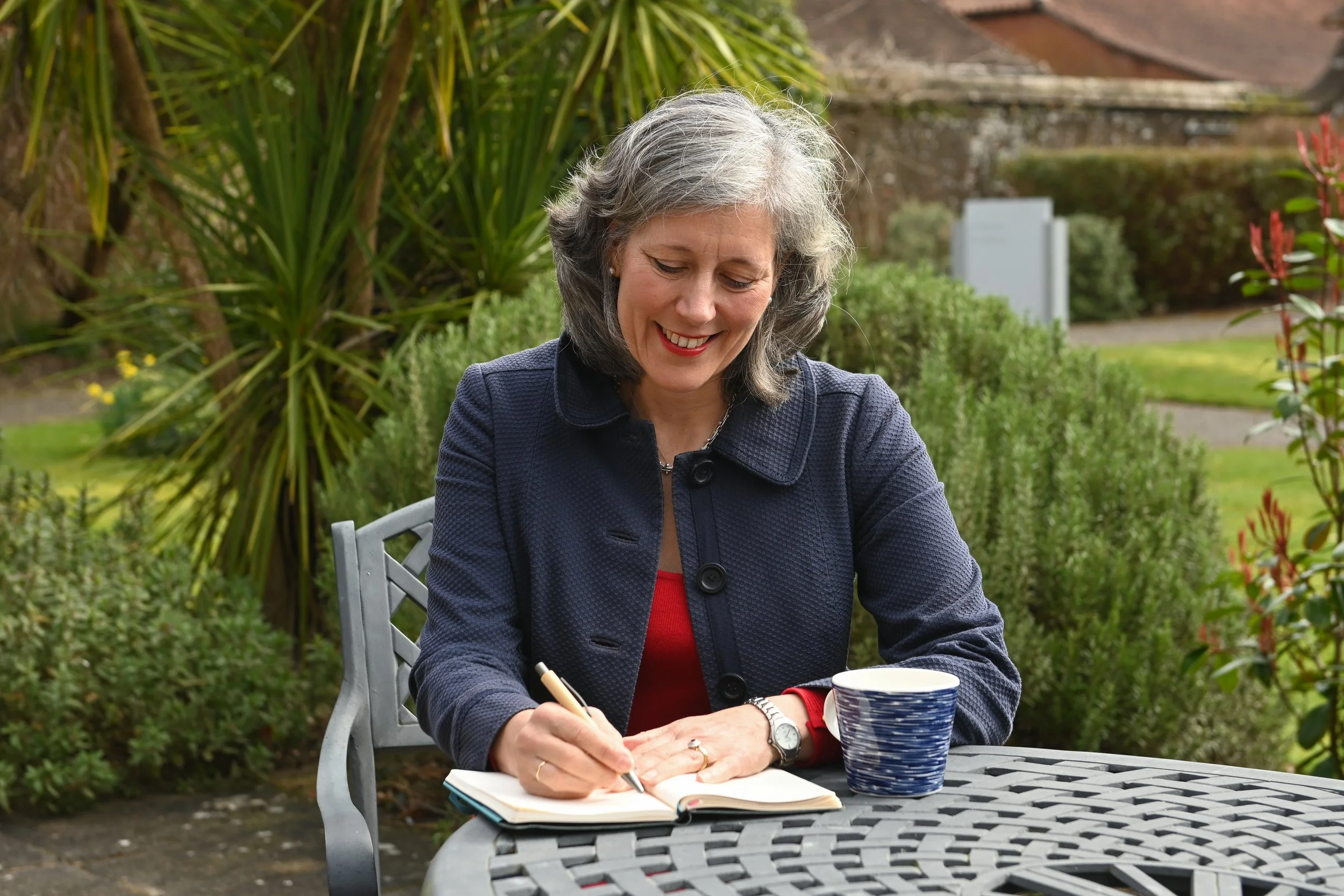 A woman with gray hair sitting at an outdoor table writing in a notebook with a pen, smiling. She has a blue and white striped mug and a cup on the table, in a garden setting with green bushes and trees.