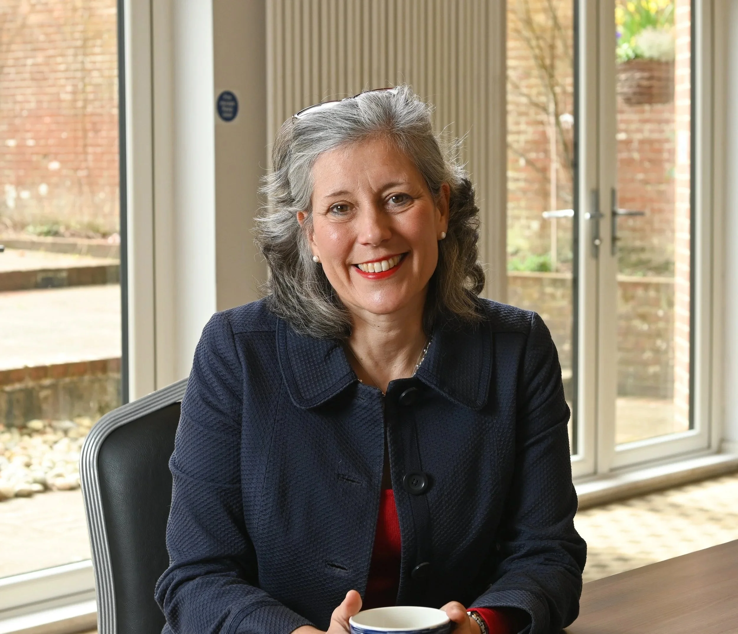 A woman with gray hair and pearl earrings sitting indoors near large windows, holding a white cup with a blue pattern, smiling at the camera.