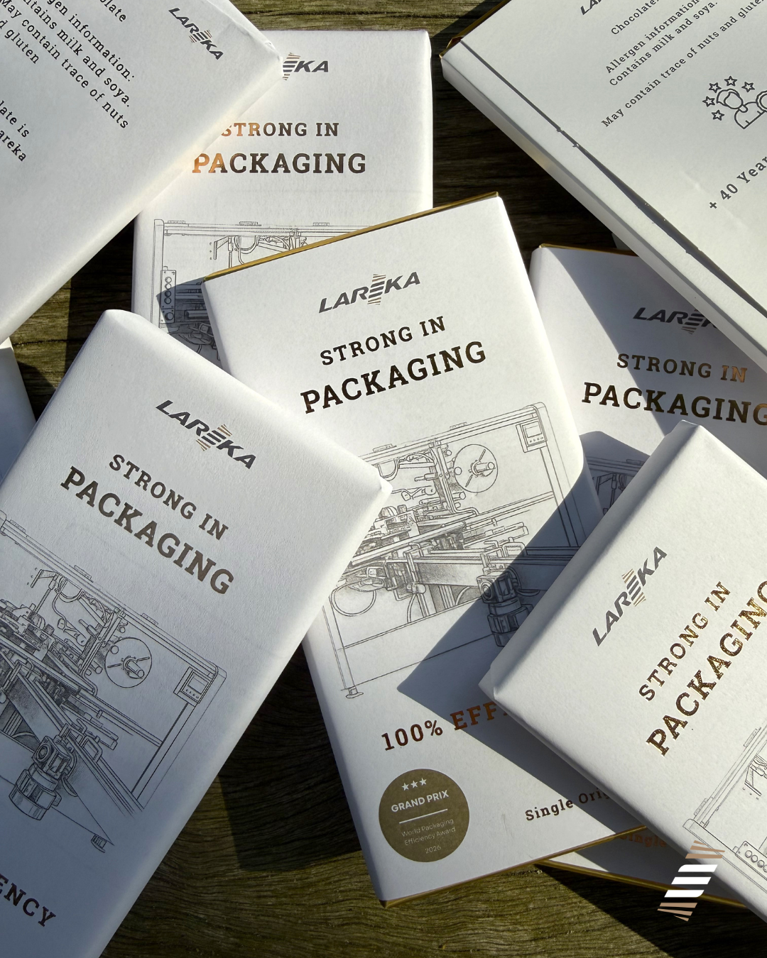 Several chocolate bars wrapped in a white paper outer layer in with gold trim and text reading "LAREKA" and "Strong in Packaging" on a wooden surface. The bars feature illustrations of the BTB25 chocolate packaging machines.