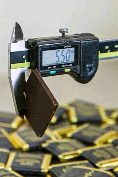 A digital caliper measuring a chocolate bar, displaying a thickness of 5.50 millimeters. Multiple packaged chocolate bars are stacked in the background.