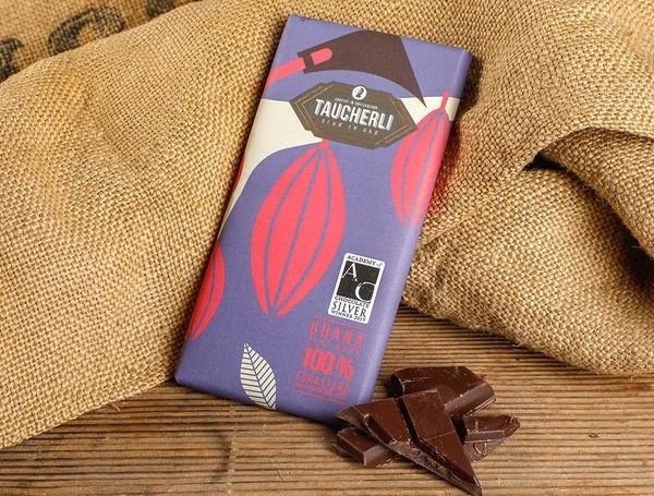 A purple chocolate bar wrapper with red and white illustrations of cacao pods, next to broken pieces of dark chocolate on a wooden surface with burlap fabric in the background.