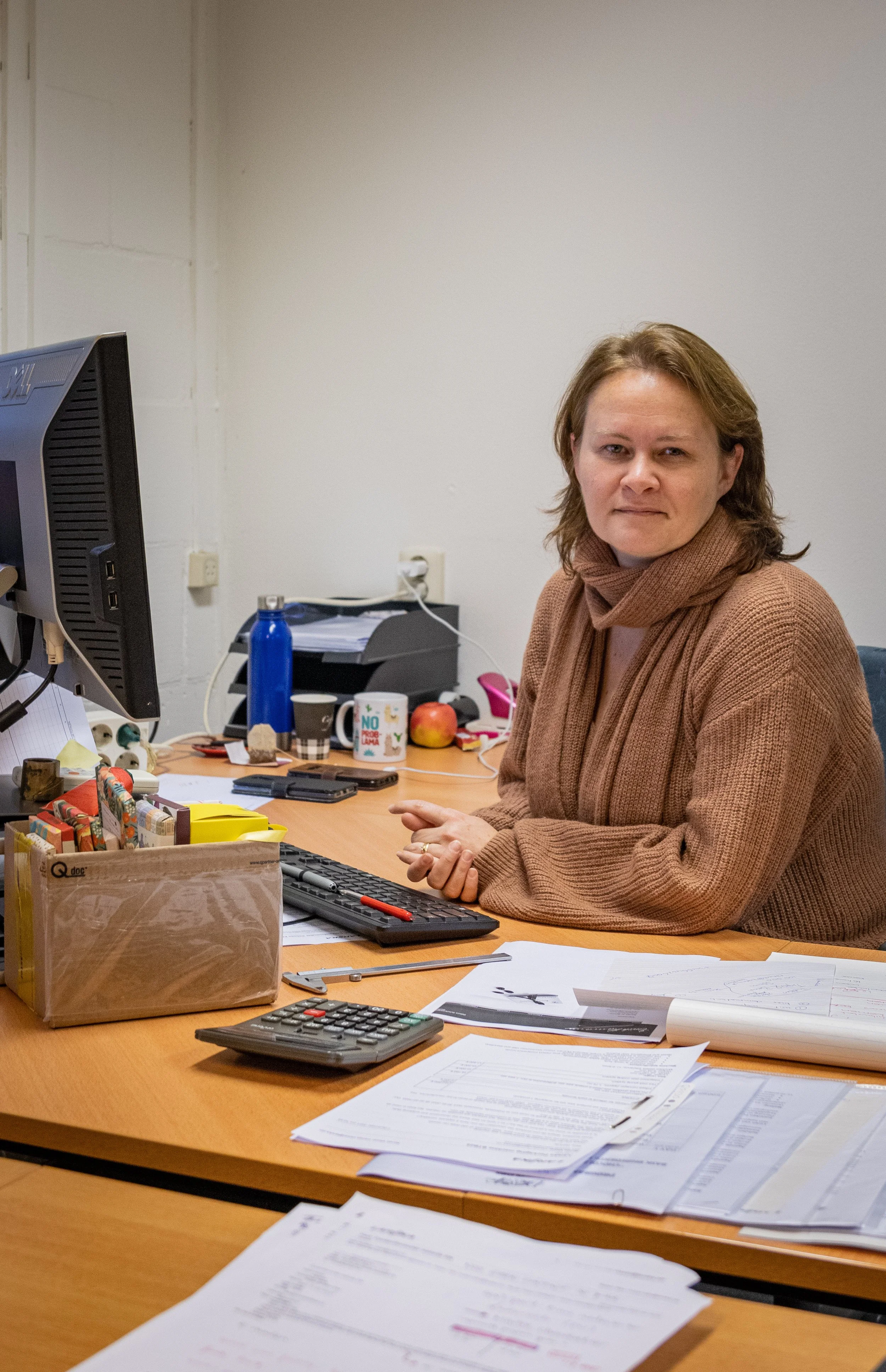 A woman from the Lareka sales team sitting at an office desk with papers, a calculator, a computer monitor, and various office supplies, looking at the camera with a slight smile.