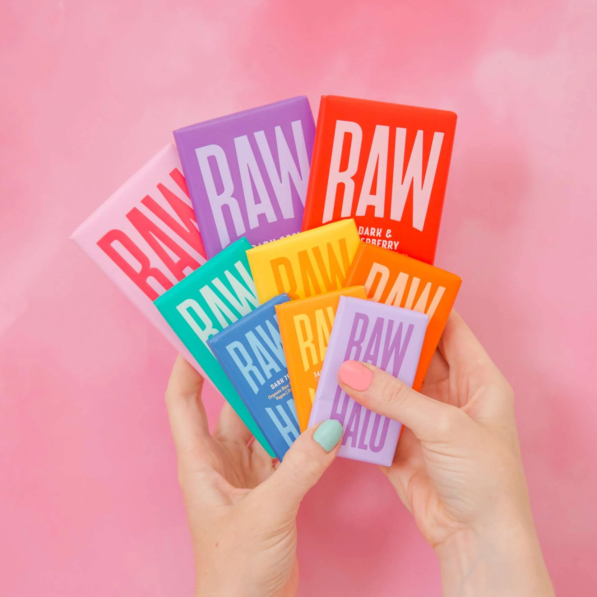 Hand holding a colorful assortment of vegan chocolate bars from Raw Halo against a pink background.