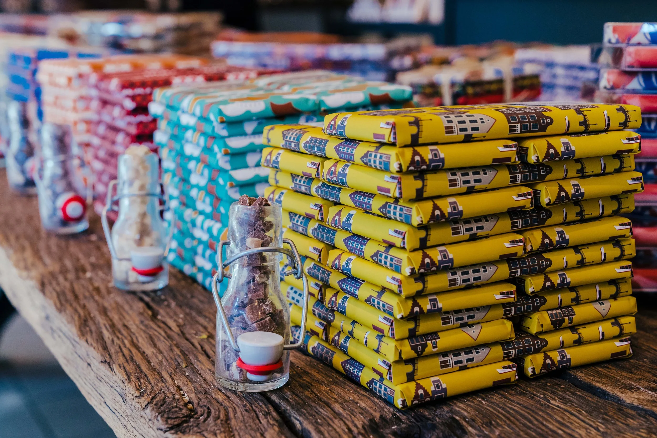 Stacks of colorful wrapped chocolate bars from Urban Cacao and small jars with candies on a wooden surface.