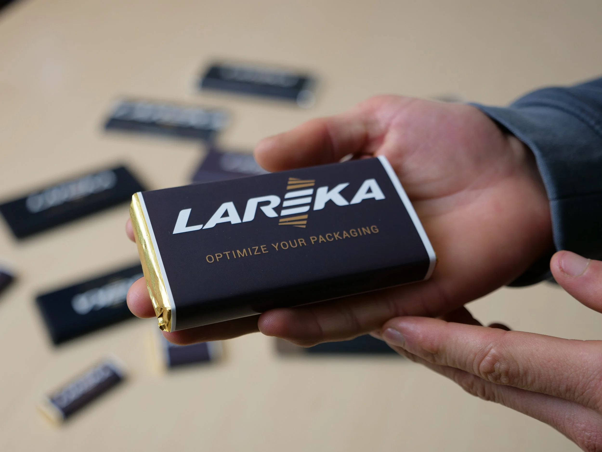 A hand holding a chocolate bar wrapped in a band wrap packaging style labeled 'LAREKA' with gold edges, with several more similarly packaged items on a beige surface in the background.