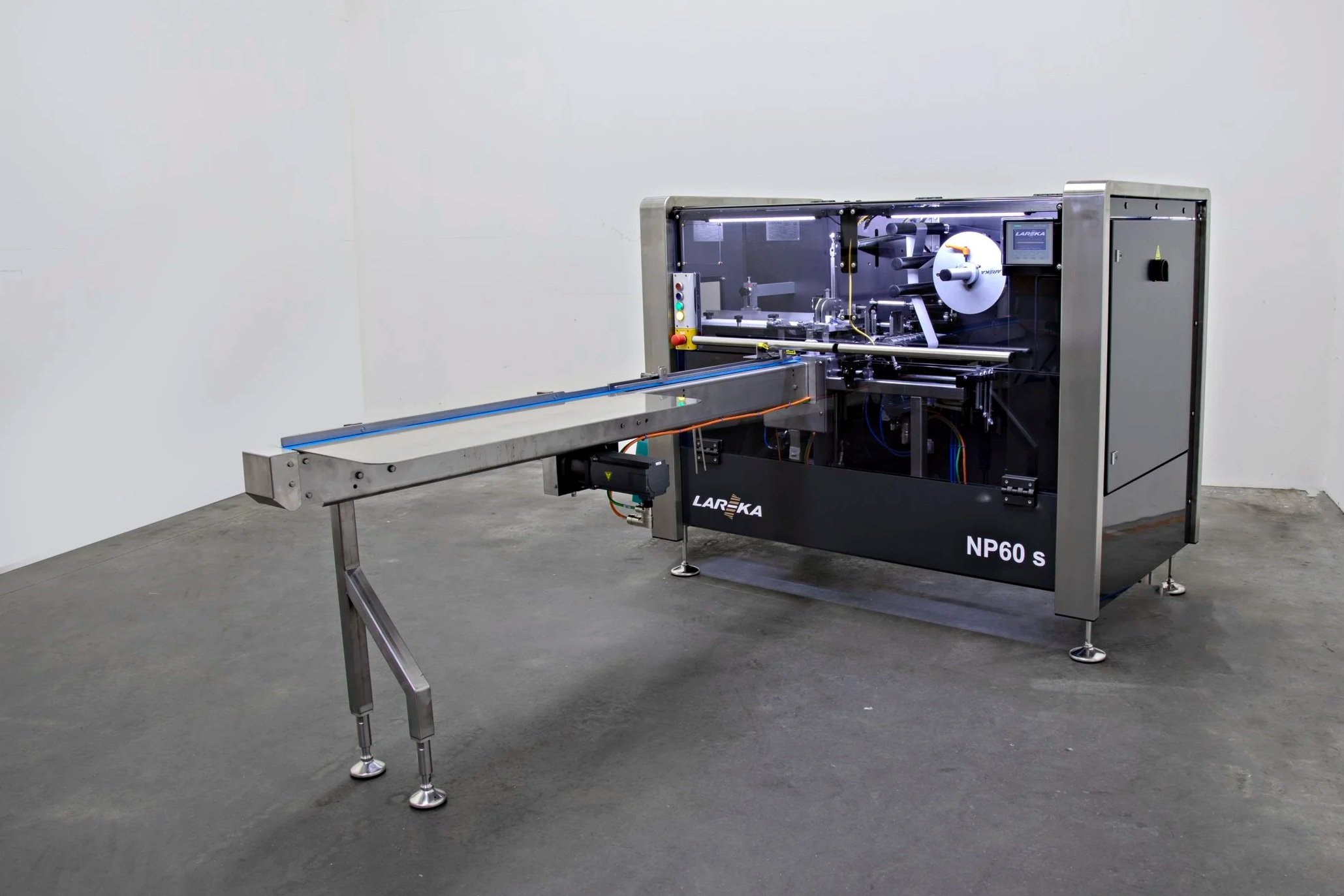 The NP60 chocolate packaging machine with a conveyor belt extending from it, placed against a plain white wall in a room with gray flooring.