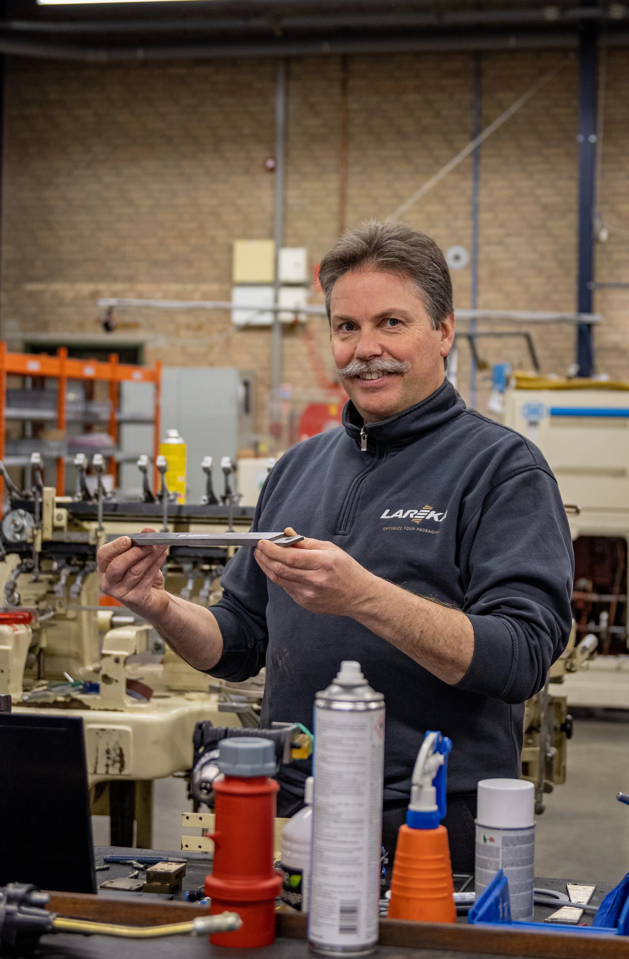 A man from the Lareka team with a mustache and gray hair smiling in a warehouse, holding a metal object, surrounded by tools with equipment and brick wall in the background.