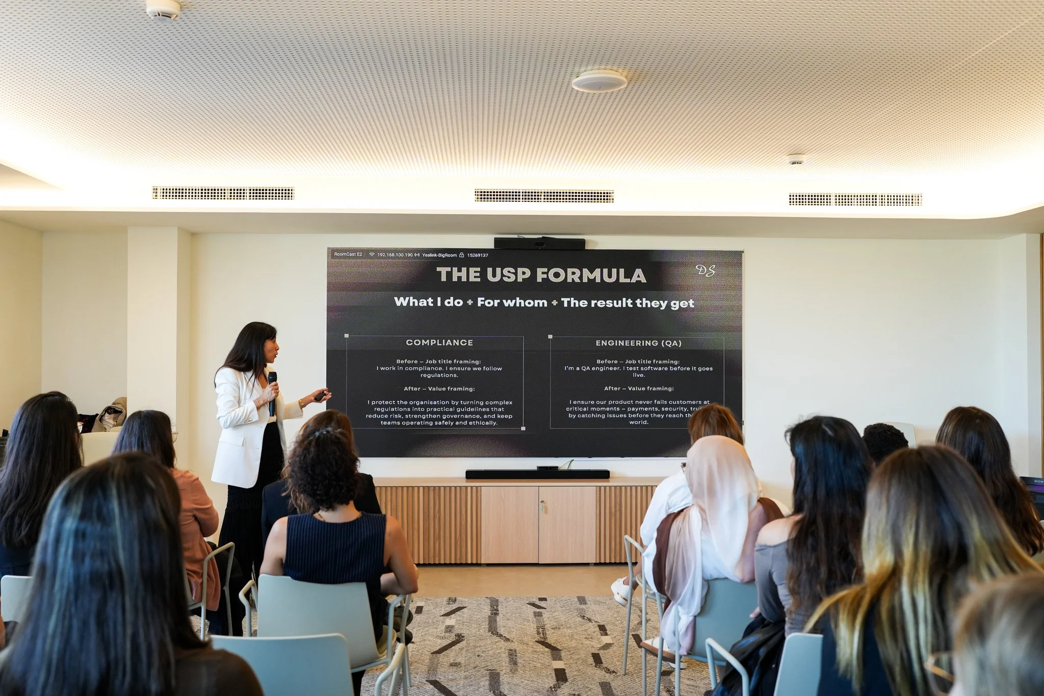 A woman in a white blazer giving a presentation to an audience in a conference room with a large screen. The screen displays a slide titled 'THE USP FORMULA' with bullet points about compliance and engineering.