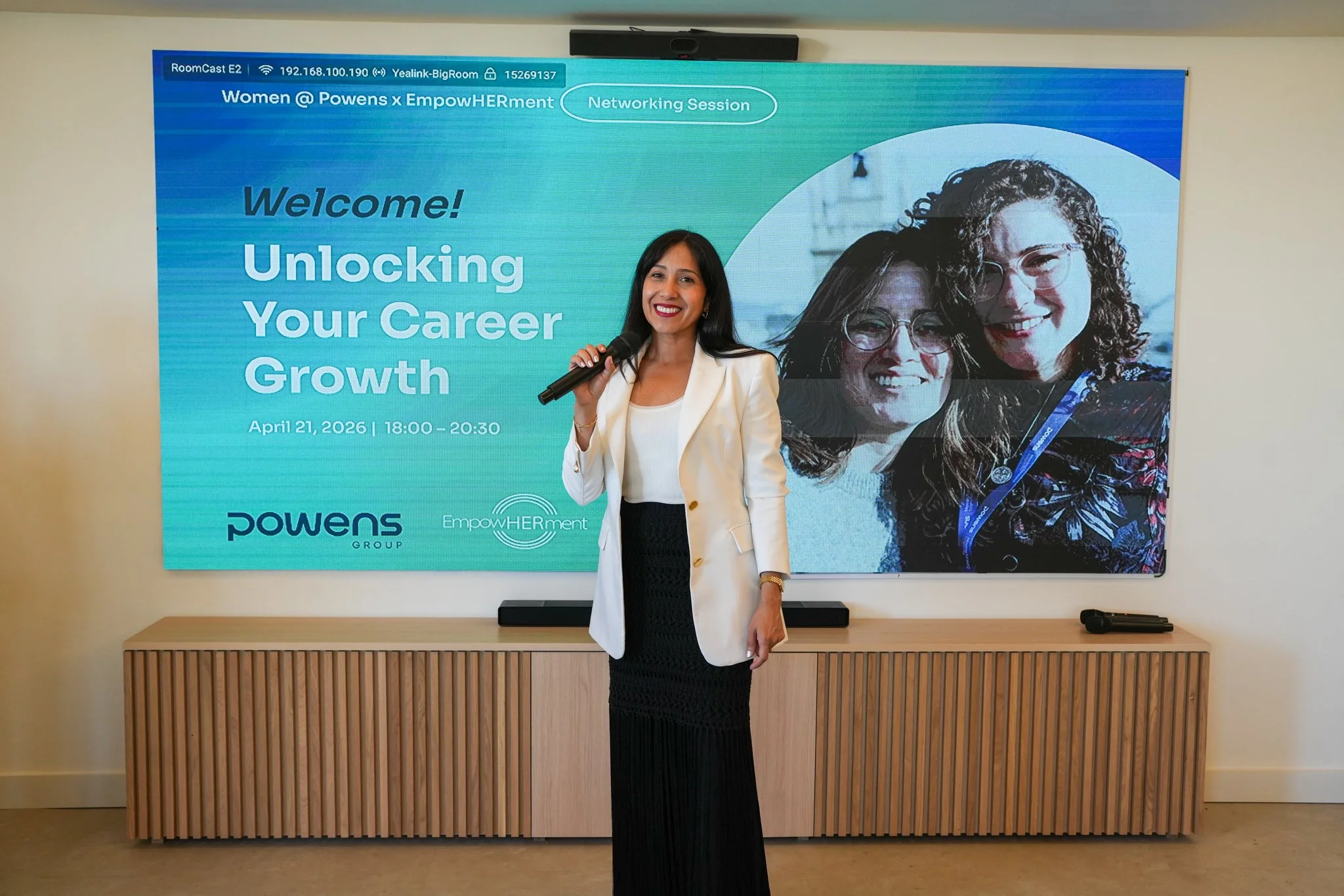 A woman in a white blazer and black dress holding a microphone stands in front of a large blue digital display at a networking event. The display reads 'Women @ Powens x EmpowHERment,' welcoming attendees to an event titled 'Unlocking Your Career Gro