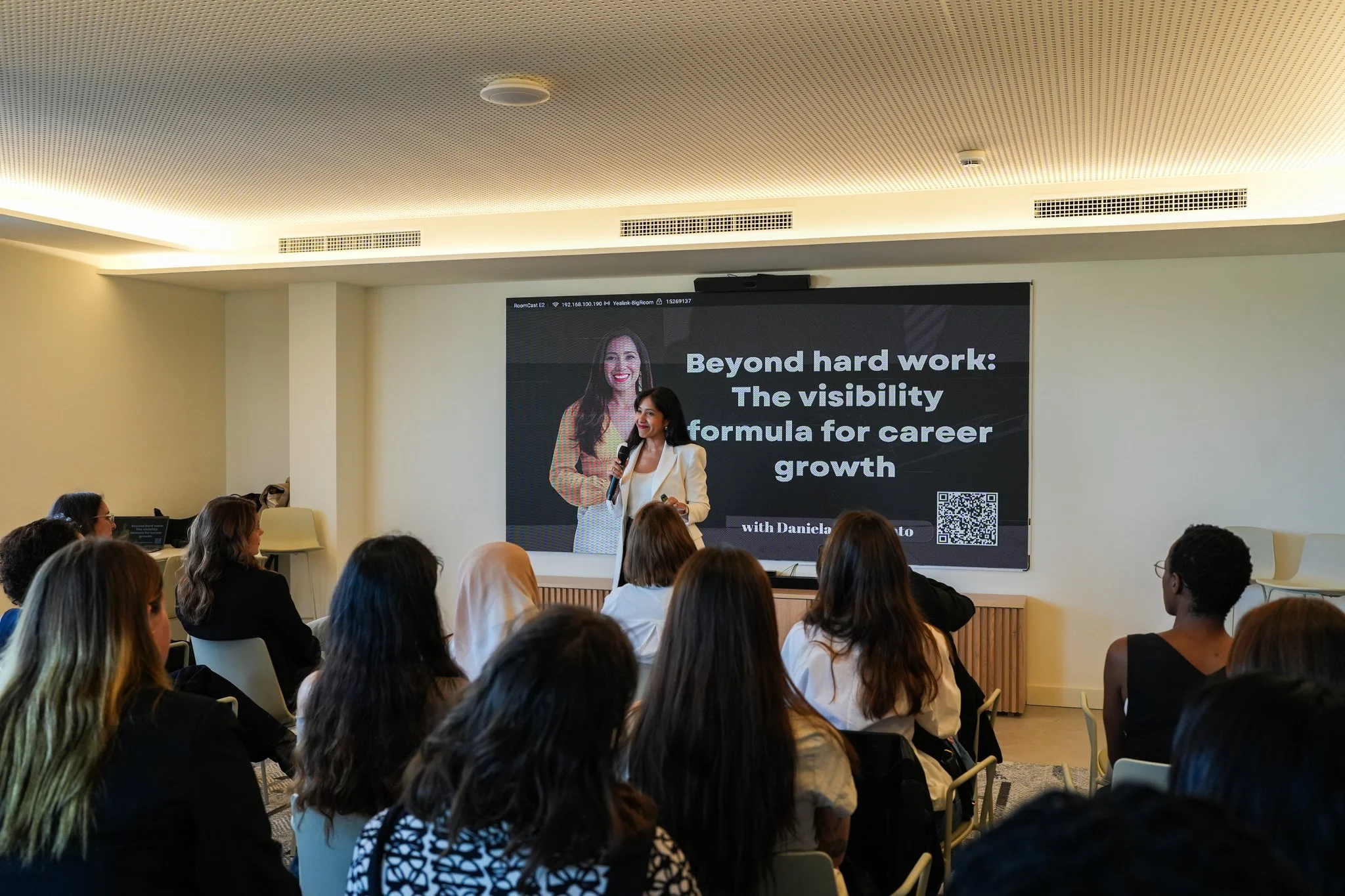 A woman presenter standing in front of a large screen at a conference, with an audience of attendees seated and listening.