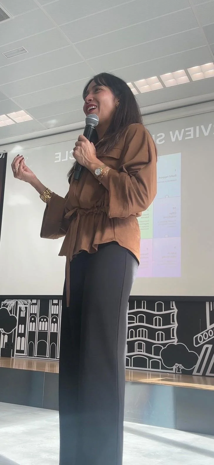 A woman speaking into a microphone during a presentation or speech, standing on a stage with a projection screen behind her and black and white cityscape illustrations on the wall.