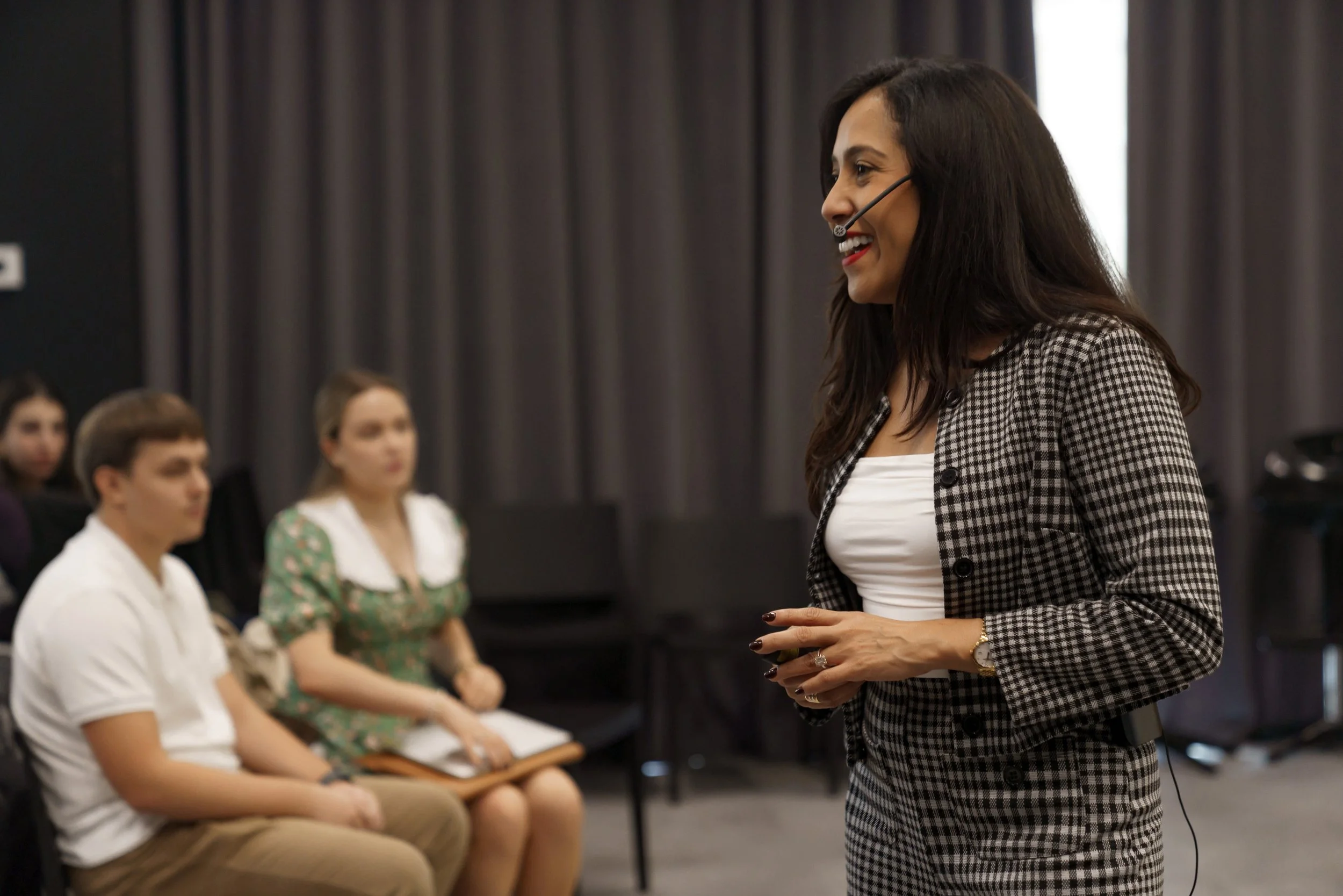 A woman with long dark hair, wearing a checkered blazer and white top, smiling and speaking during a presentation or seminar. She is using a headset microphone. In the background, some individuals are seated, attentively listening, with a dark curtai